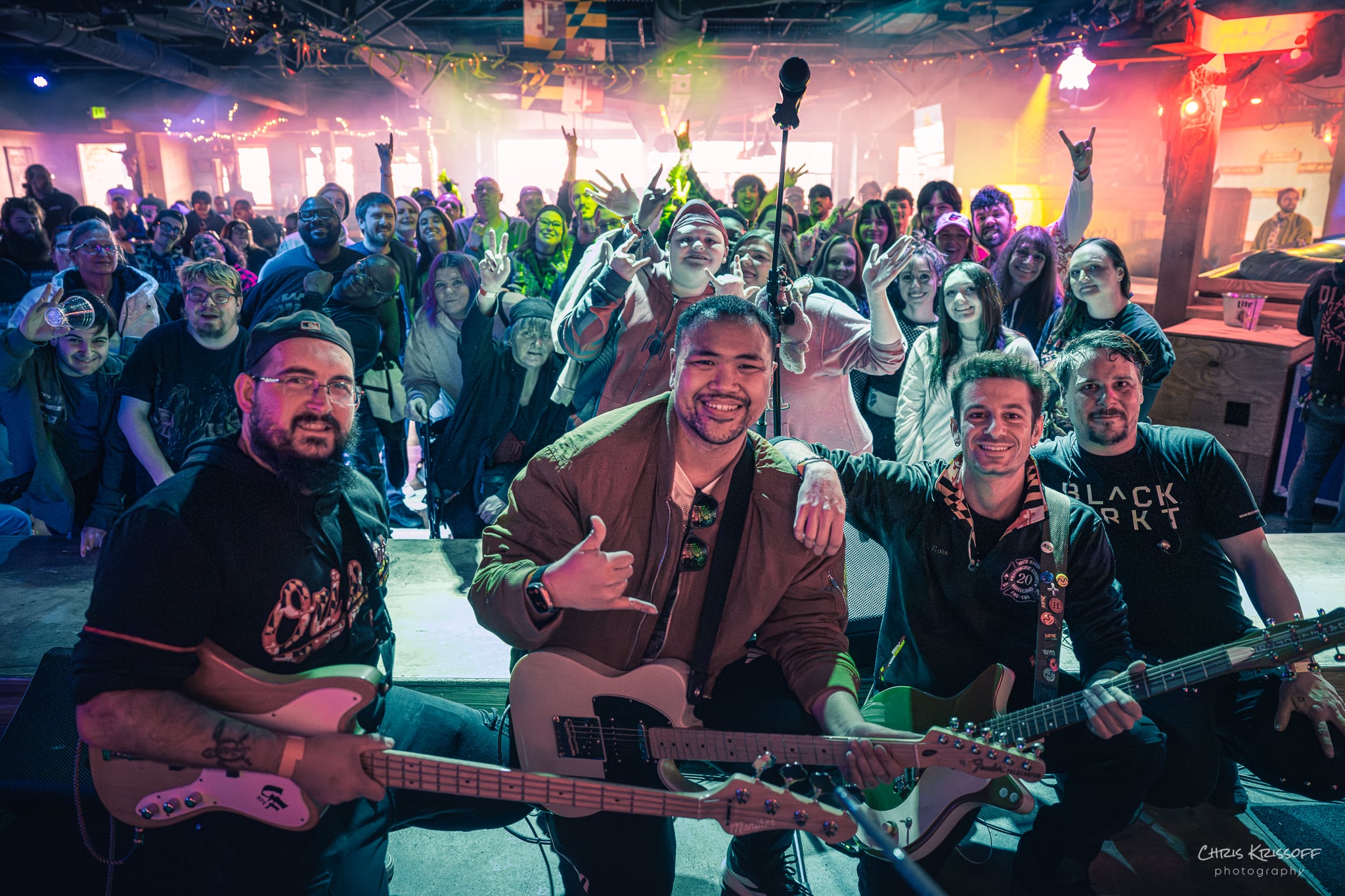Four musicians with guitars perform on stage in front of a crowd at a concert, colorful lights illuminate the scene.