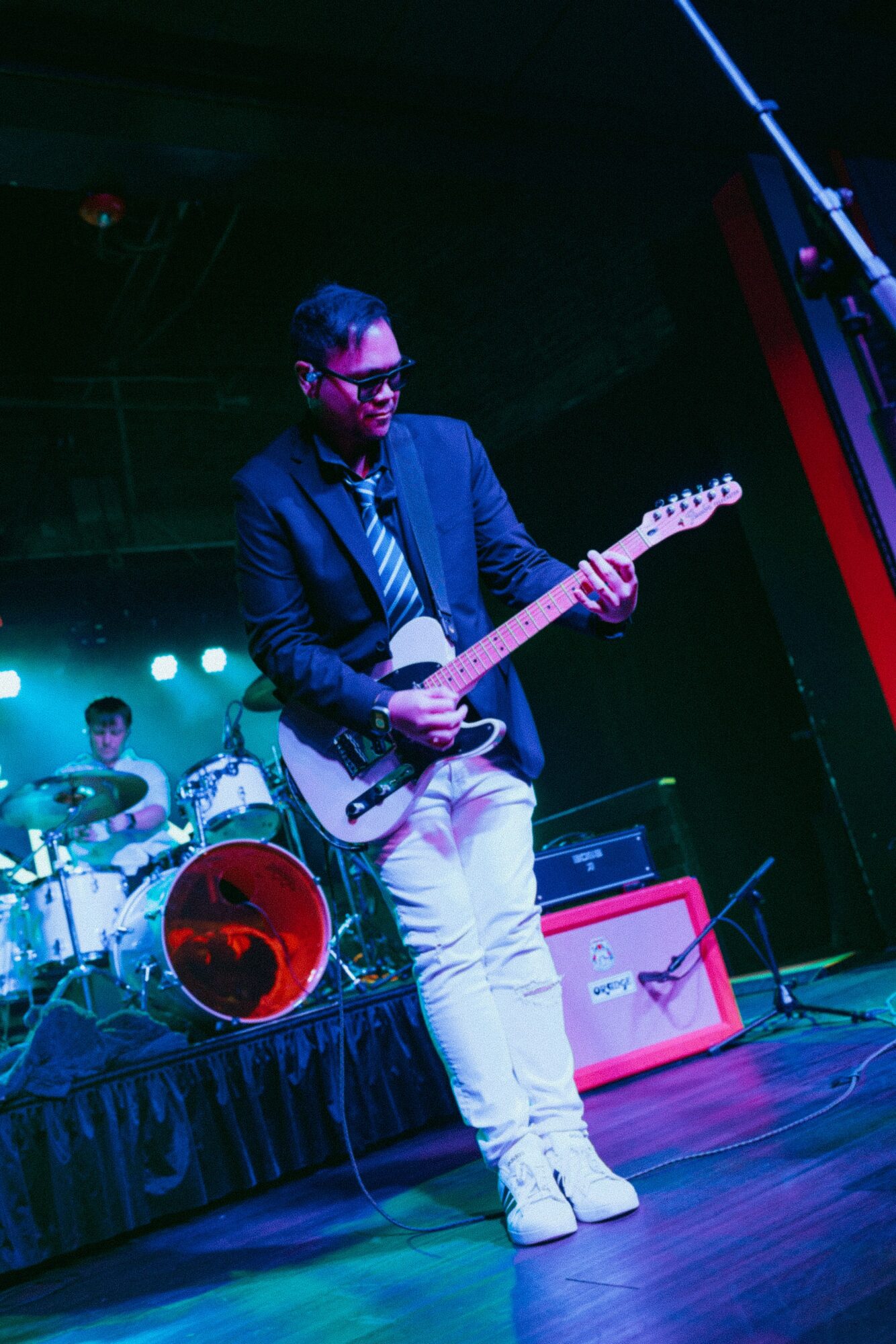 Musician playing electric guitar on stage with drummer in background, colorful stage lighting, and musical equipment visible.