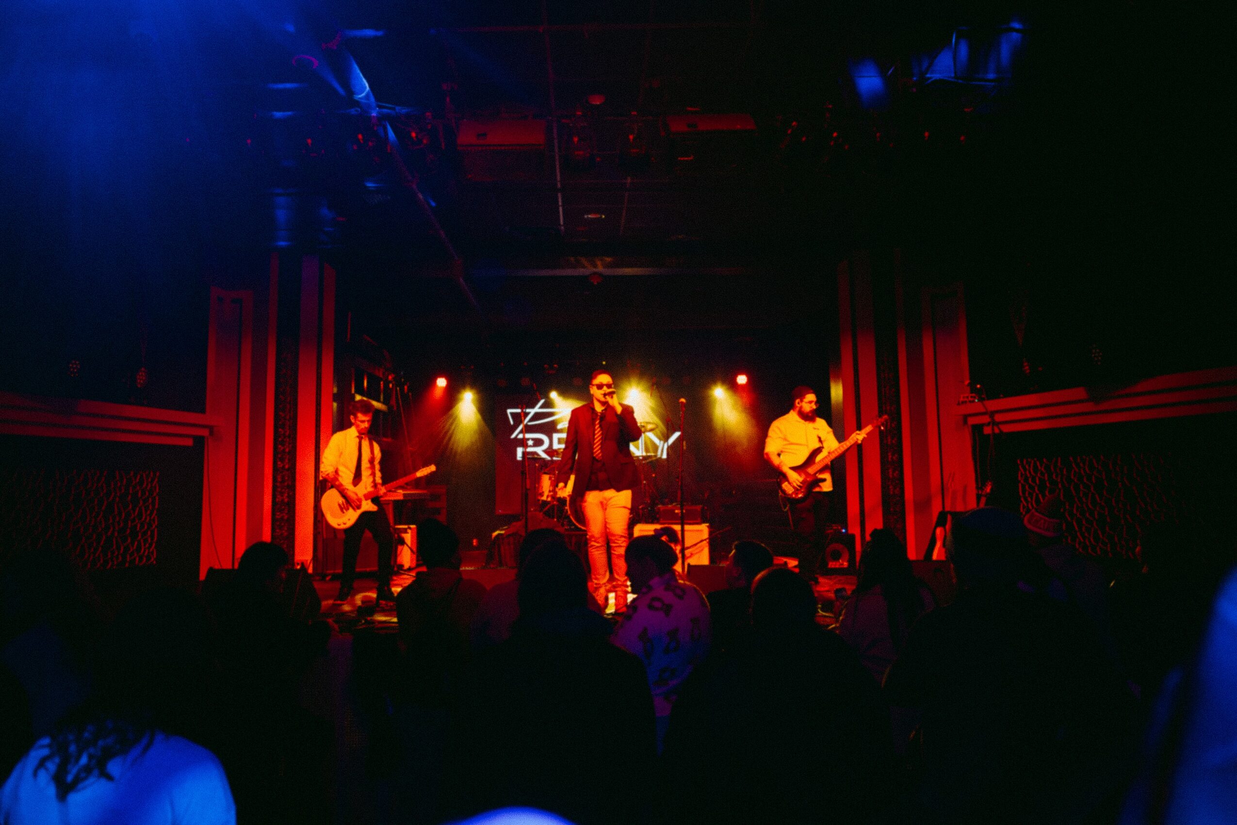 Band performs on stage with red curtains, bright lights, and audience in foreground, in a dark indoor venue.