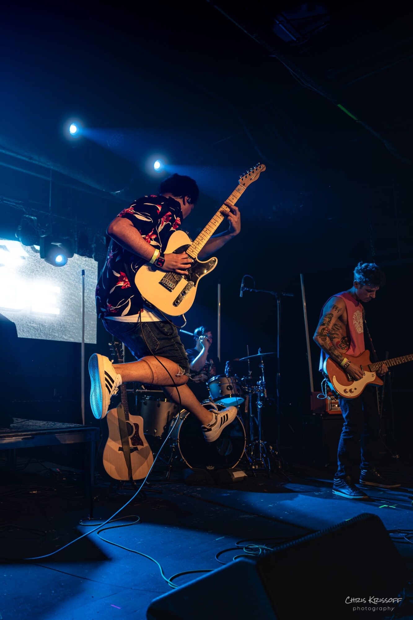 Musicians perform on stage with guitars, illuminated by stage lights, in a dark concert setting.