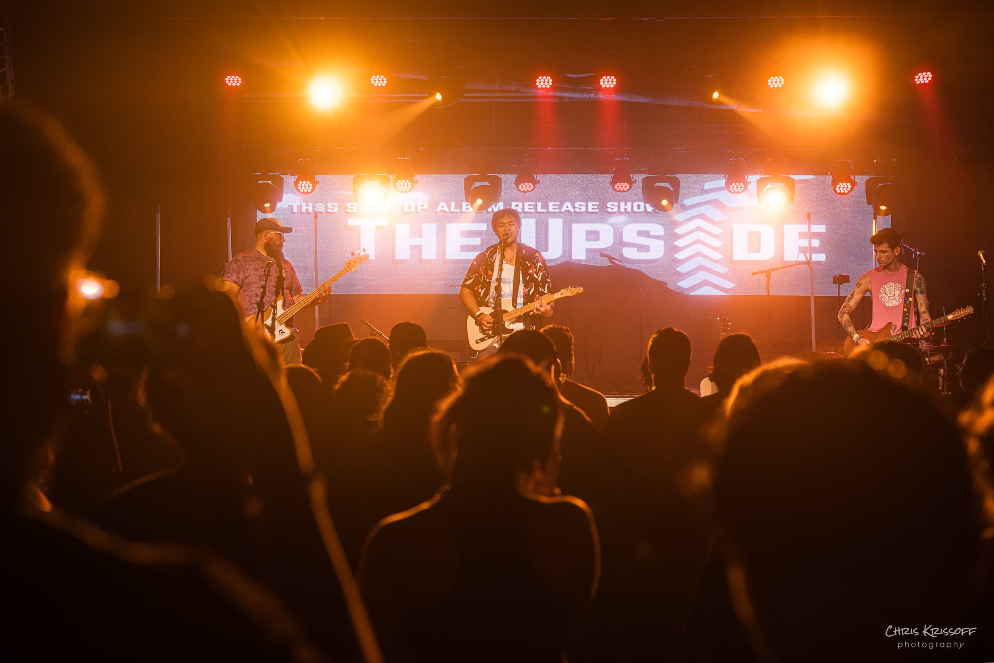 Band performs on stage with bright orange and yellow lights, audience silhouettes in foreground, large screen behind displays text.