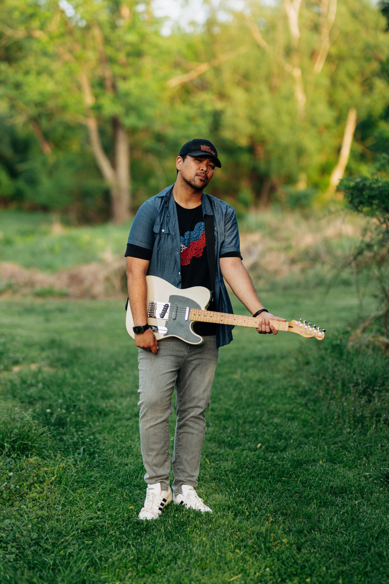 Young man standing outdoors holding an electric guitar, wearing a cap, glasses, t-shirt, and jeans, with trees in the background.