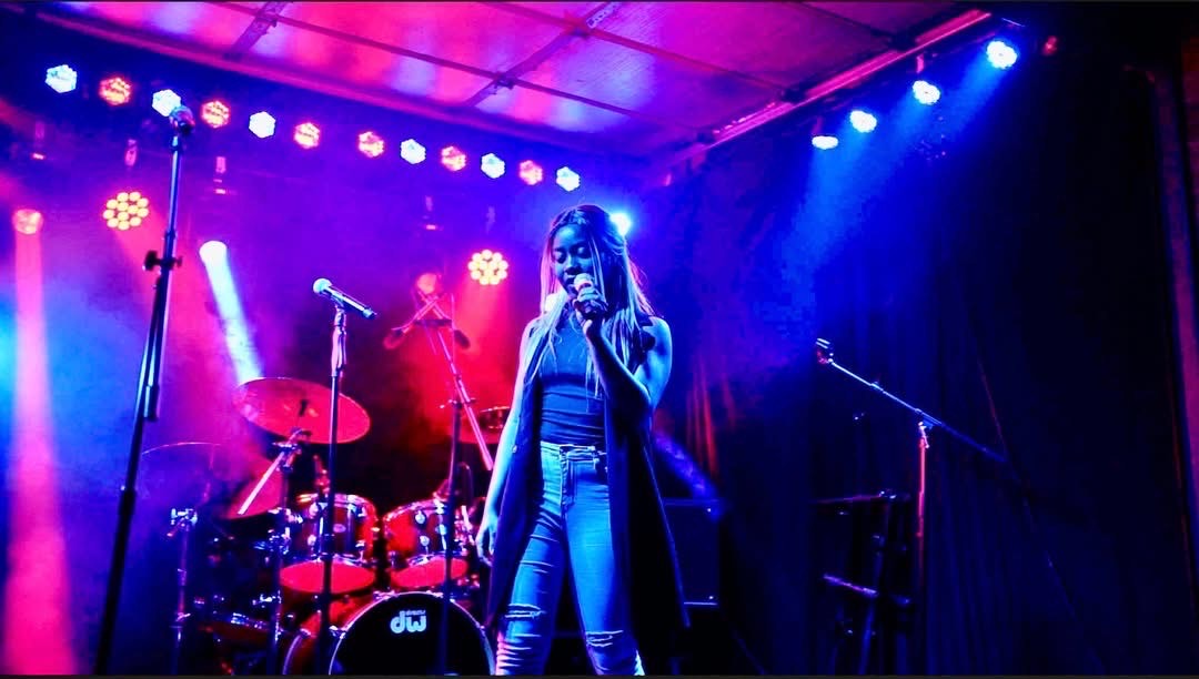 A woman stands on stage with colorful lighting, holding a microphone, with drums and microphones in the background.