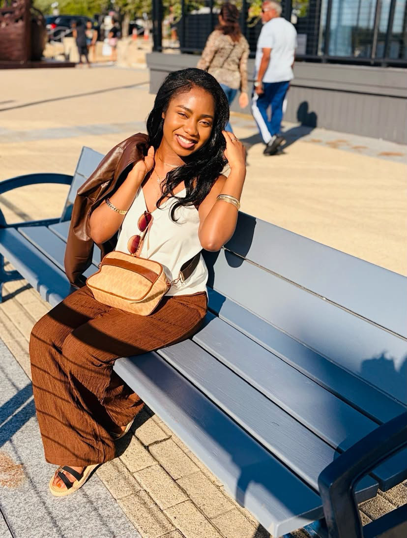 Smiling woman sitting on a bench outdoors, wearing a white top and brown pants, with a bag on her lap.