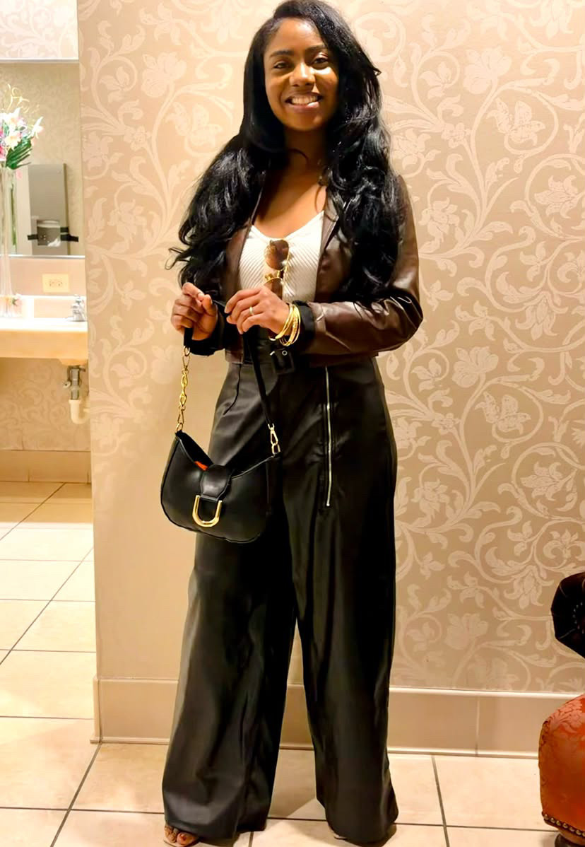 Woman with long dark hair smiling, wearing a black leather jacket, white top, and black wide-leg pants, holding a small black purse, standing indoors against a patterned wall.