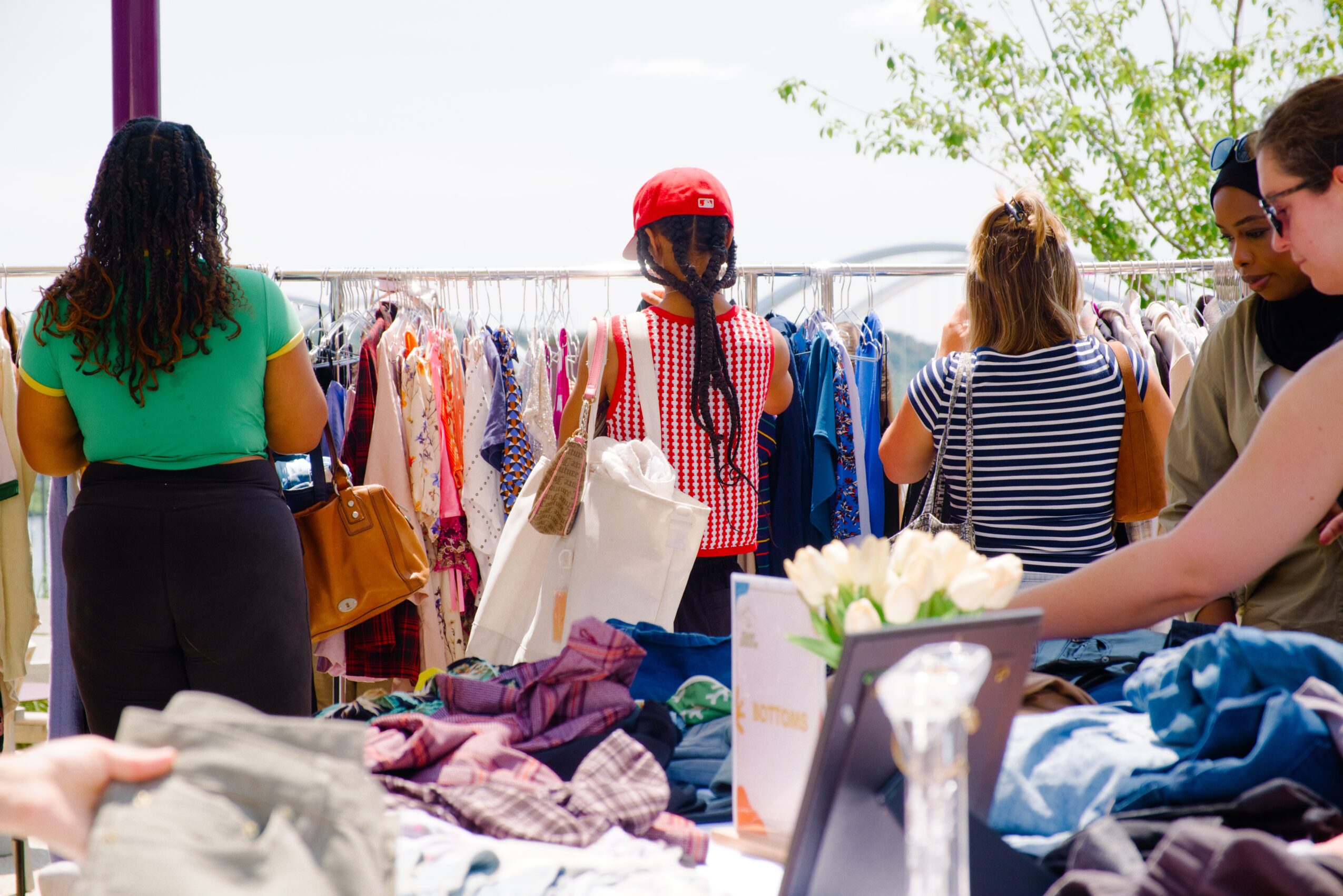 People shopping at a clothing store with racks of clothes, some browsing and others examining items, outdoors with trees in background.