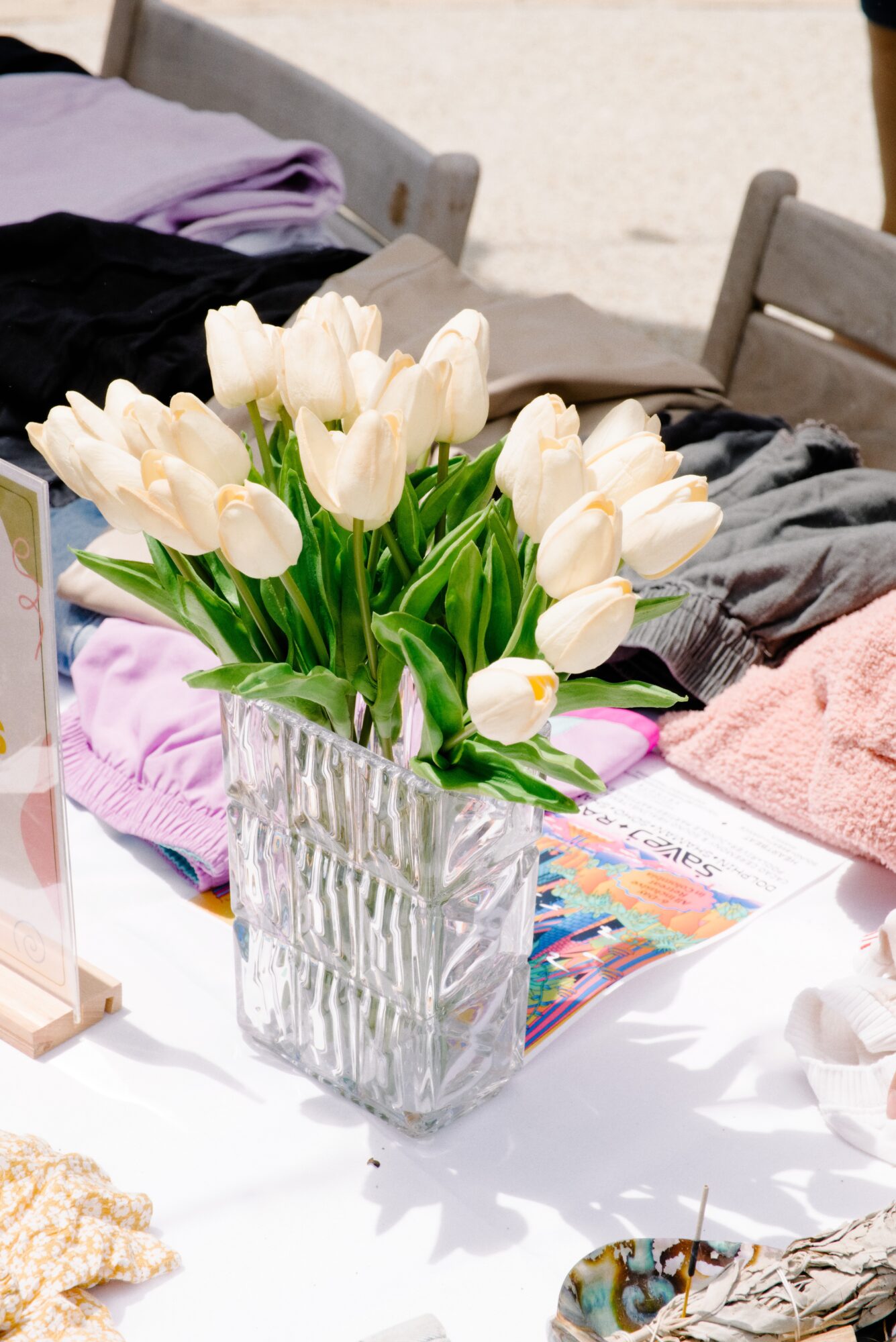 White tulips in a glass vase on a table with clothes and magazines, chairs in the background.