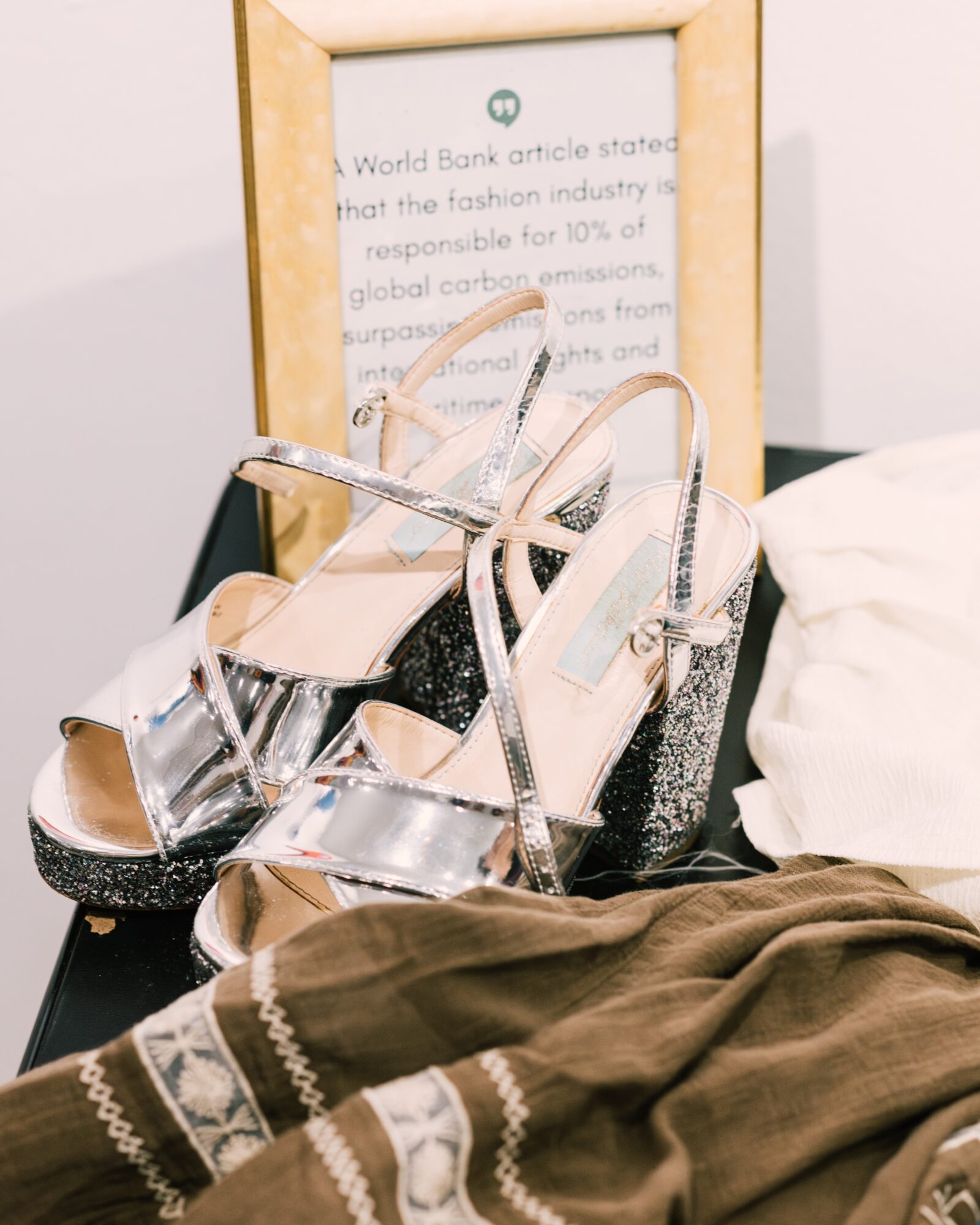 Pair of high-heeled shoes with silver straps and black glitter heels resting on a bed with brown and white bedding, framed picture in background.