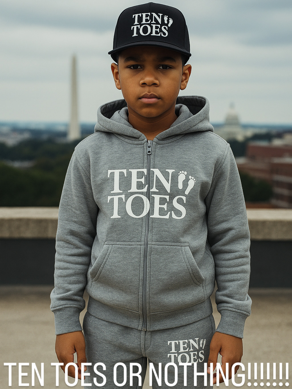 Young boy wearing a black cap and gray hoodie with 'TEN TOES' text, standing outdoors with a cityscape background.