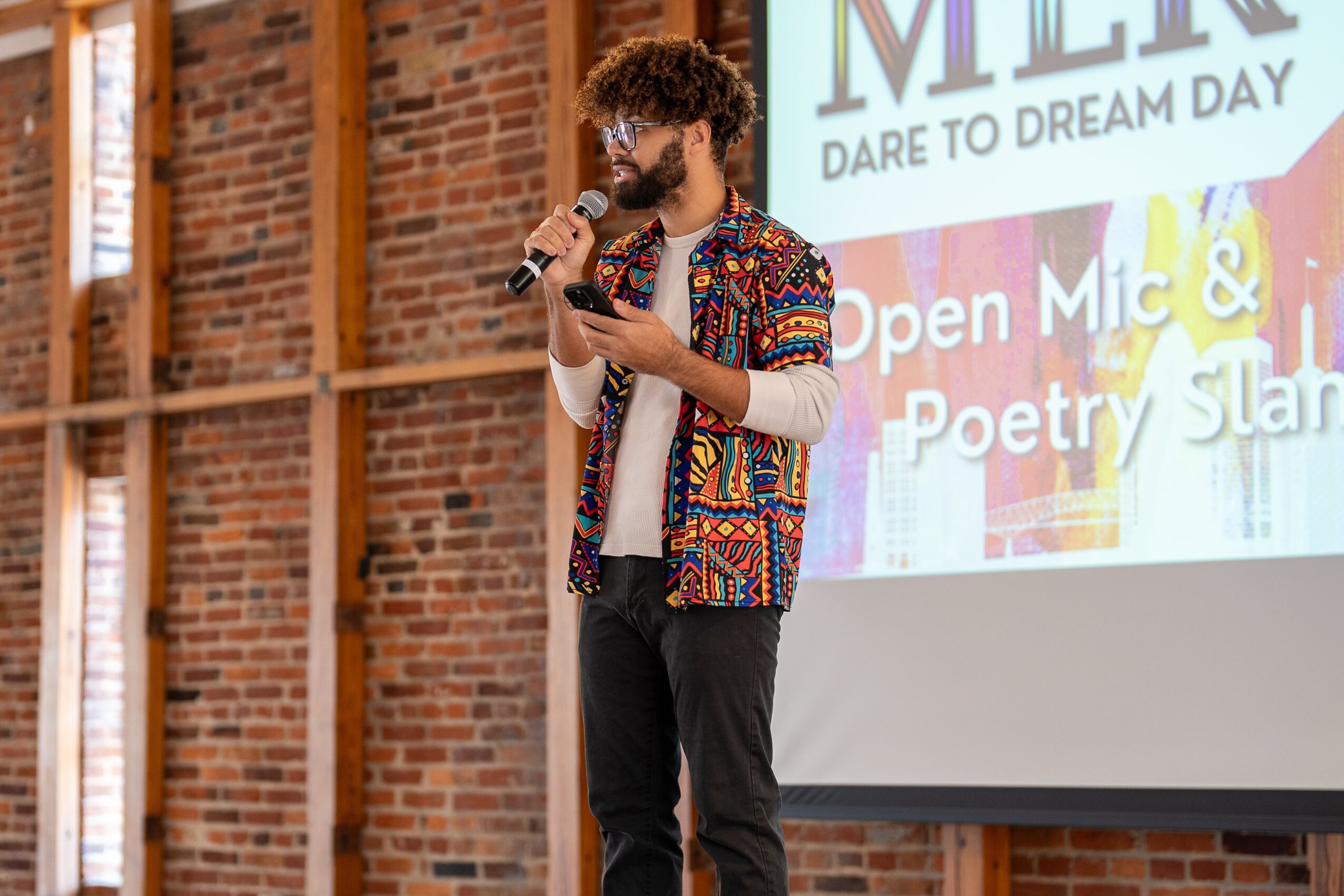 Person with curly hair and glasses speaking into a microphone on stage, brick wall background, screen with text behind.