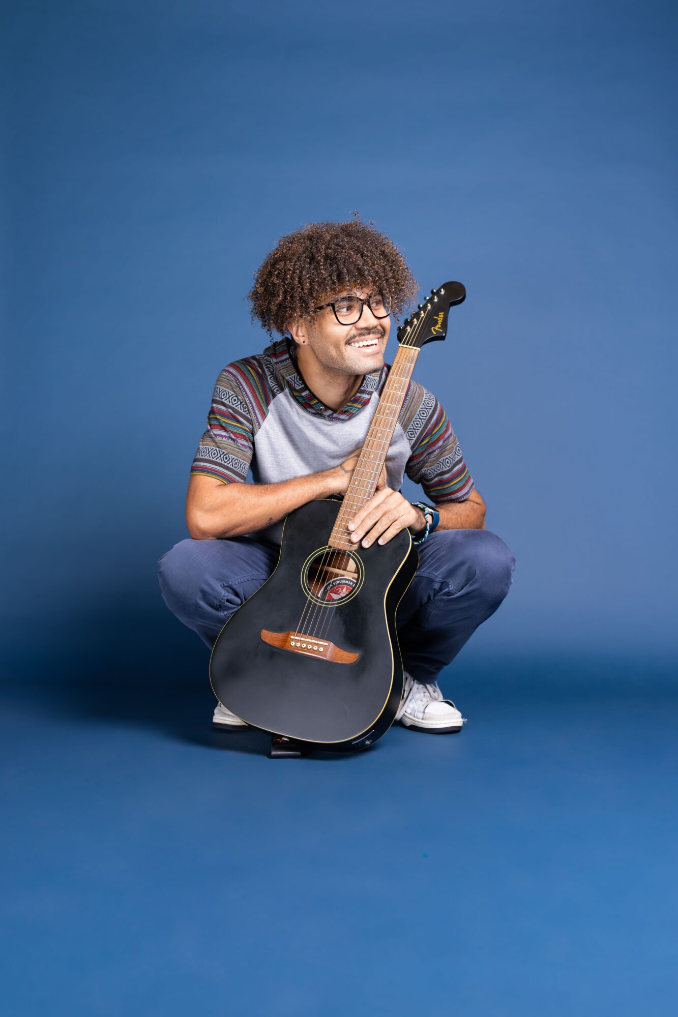 Person with curly hair and glasses crouching with a black acoustic guitar against a blue background.