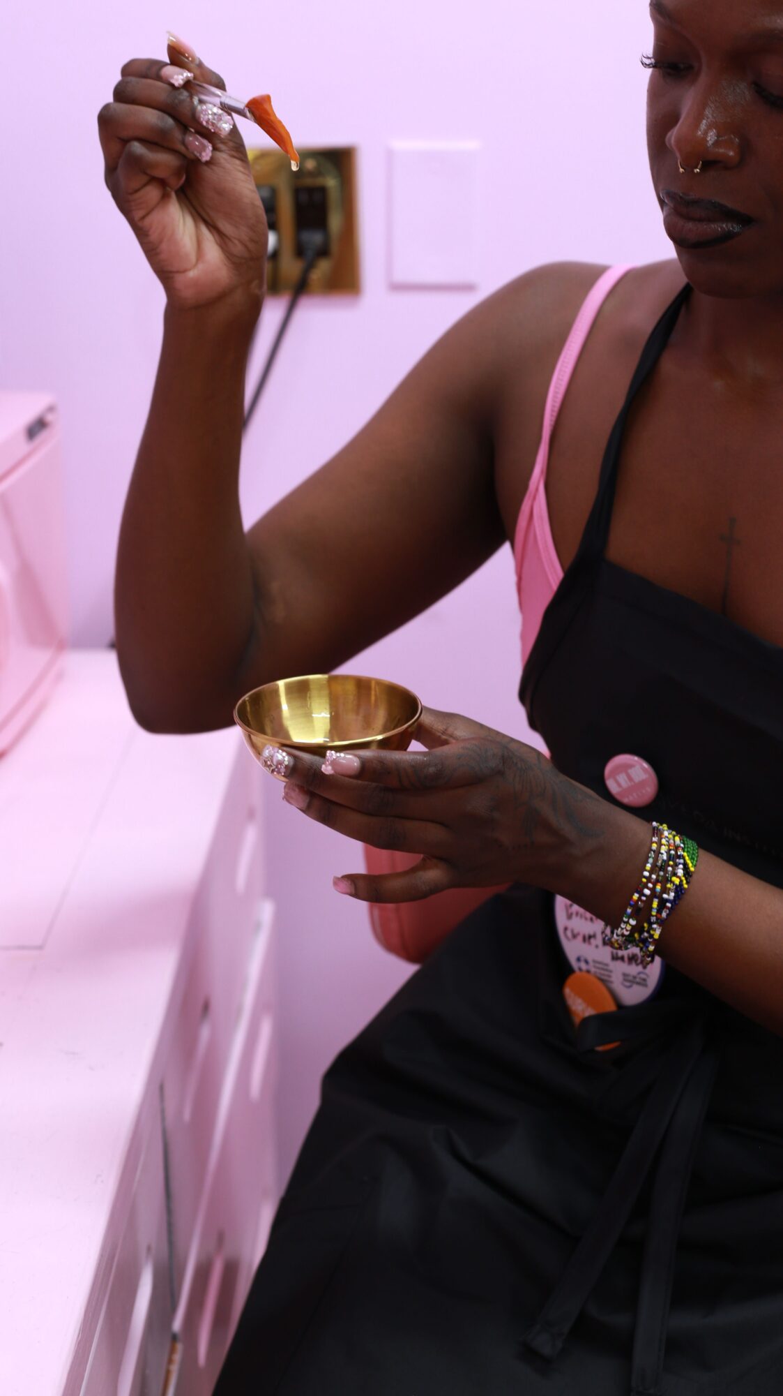 Person holding a small container, wearing a bracelet, with a pink background and a person in black clothing.