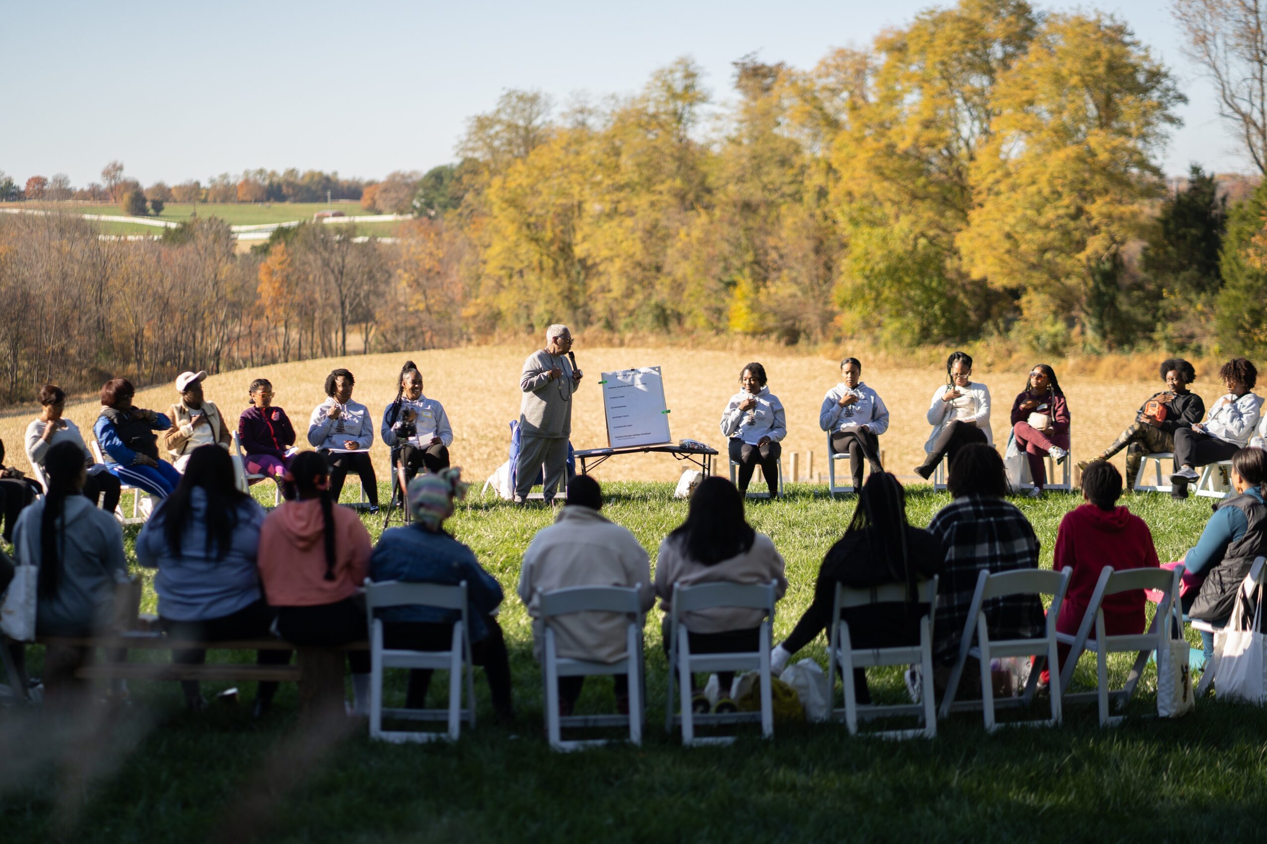 Group of people sitting outdoors in a circle, listening to a speaker with trees and autumn foliage in the background.