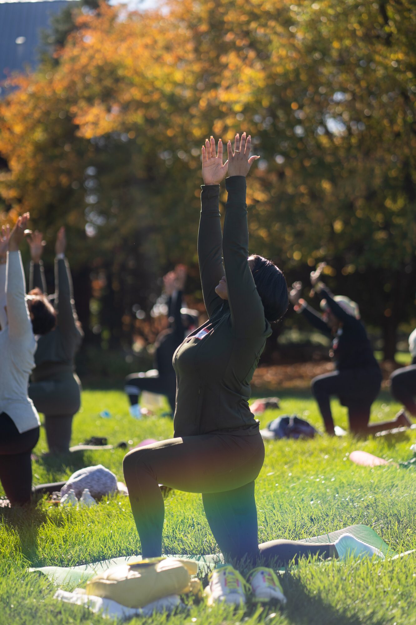 Person practicing yoga outdoors on grass with others, trees with autumn leaves in background, person in a kneeling pose with arms raised.