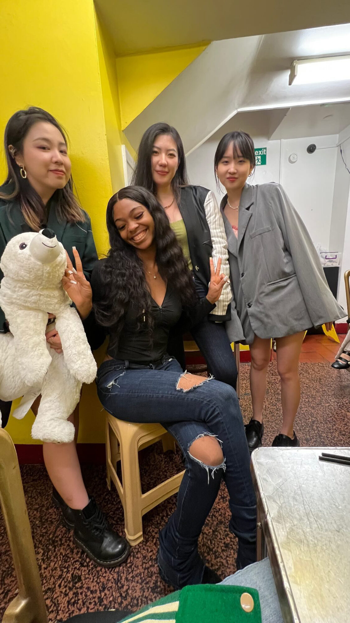 Four women posing indoors, one holding a stuffed animal, with a yellow wall and modern interior in the background.