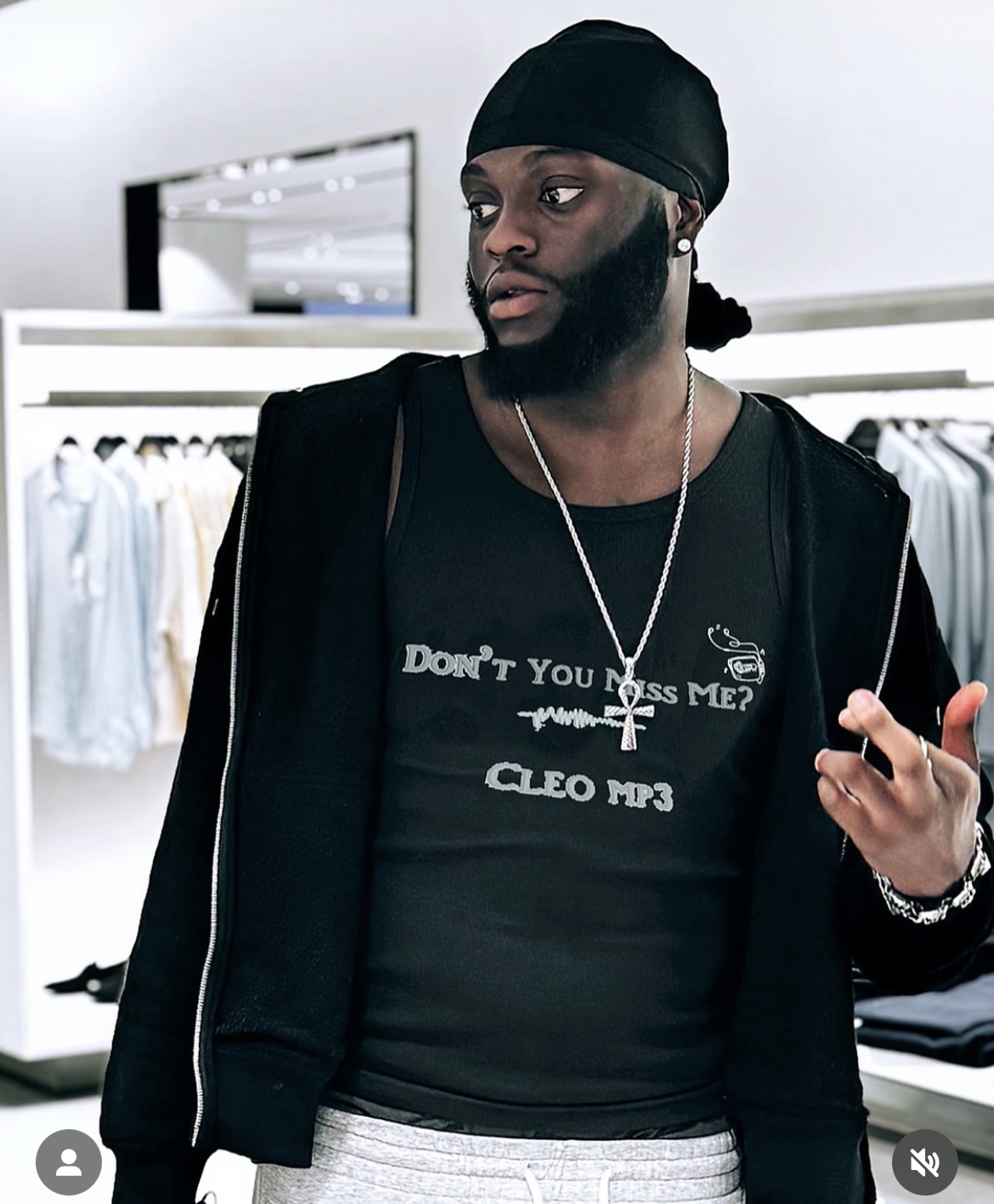 Man with a beard and black head covering in a clothing store, wearing a black T-shirt and jacket, holding a phone.