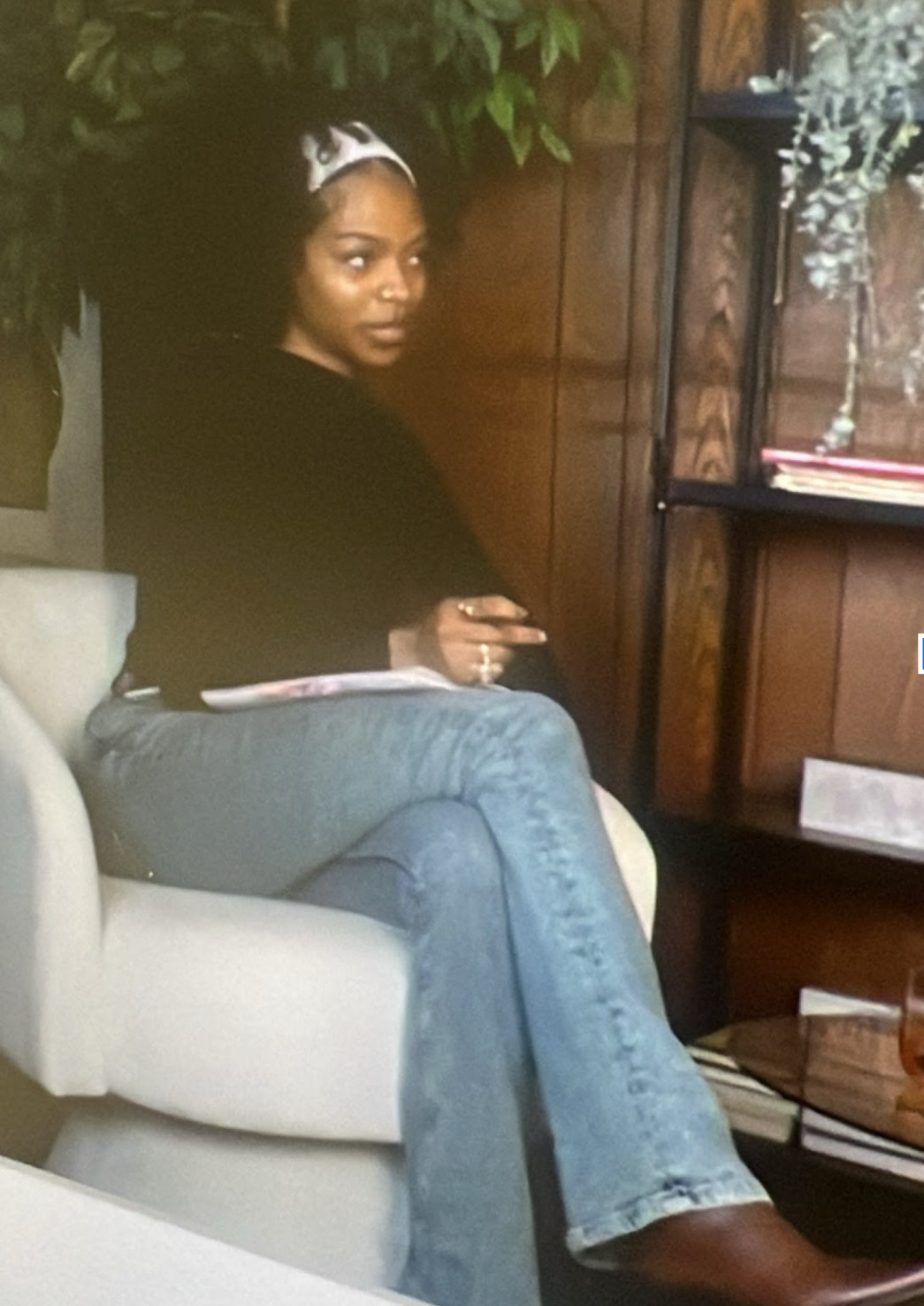 Woman sitting on a light-colored armchair, wearing a black top and jeans, in a room with wooden walls and shelves.