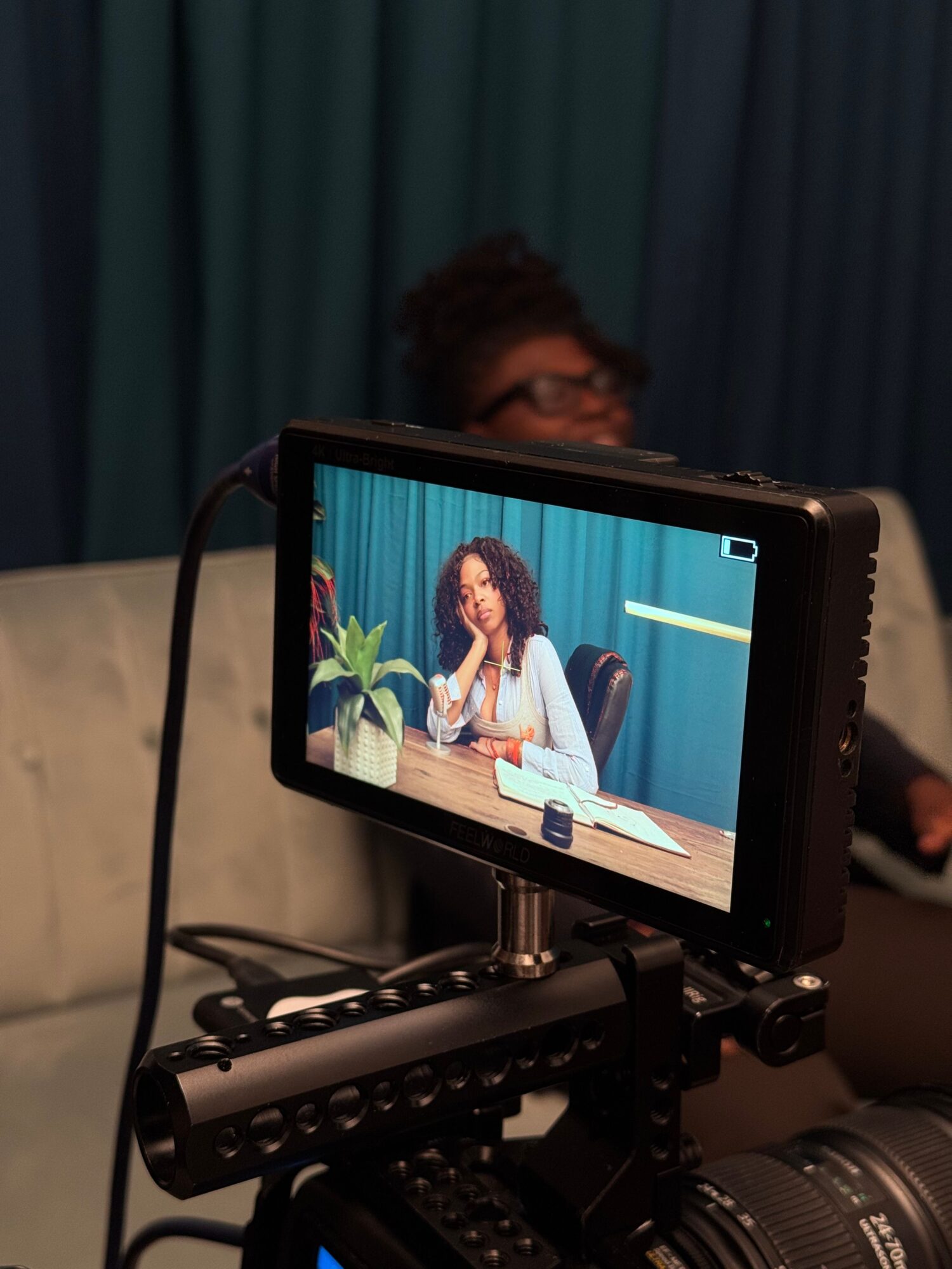 Person with curly hair and glasses sitting at a table with a plant, seen on camera monitor, background with blue curtains