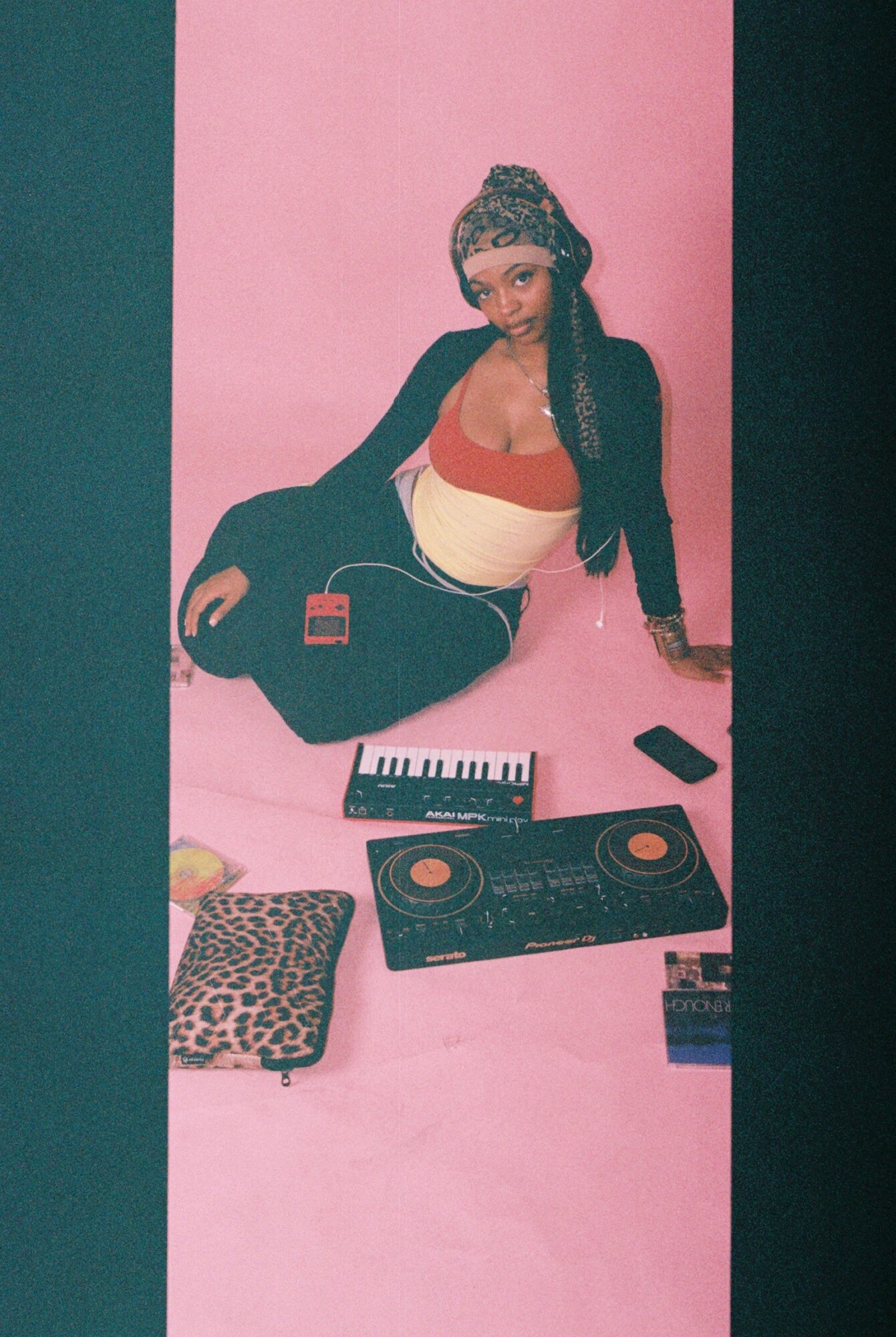 Woman sitting on pink background with music equipment, wearing a headscarf, black jacket, and colorful top.