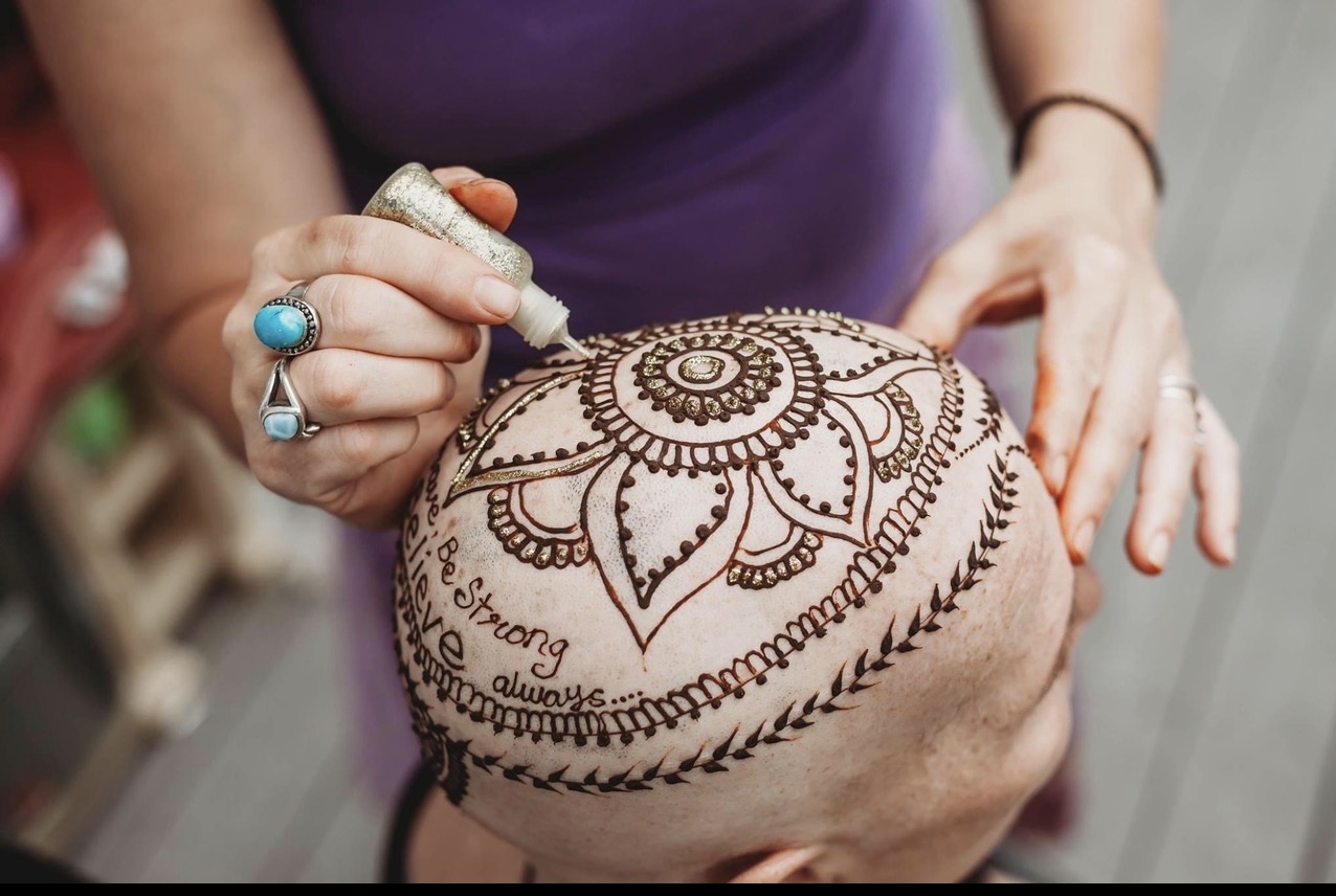 Person with a ring on finger draws intricate henna design on a bald person's head, with a purple shirt visible.