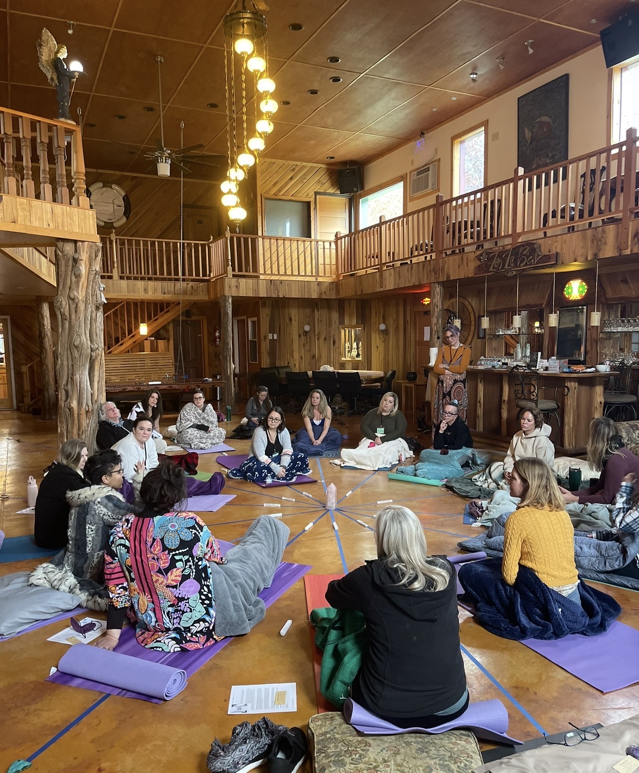 Group of people sitting on yoga mats in a wooden indoor space with high ceiling and balcony, participating in a class or workshop.