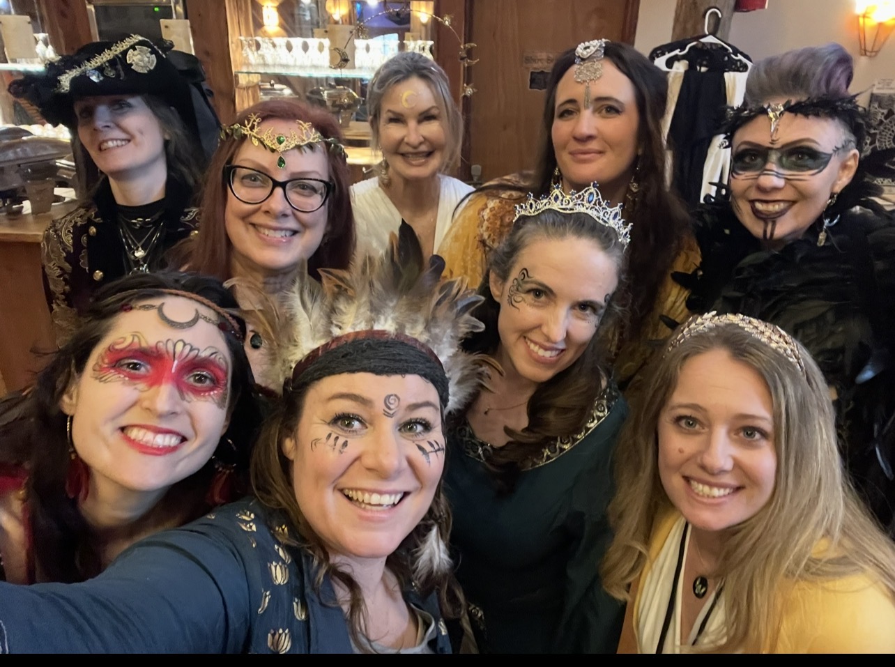 Group of women dressed in costumes and accessories, smiling indoors with wooden walls and clothing in the background.