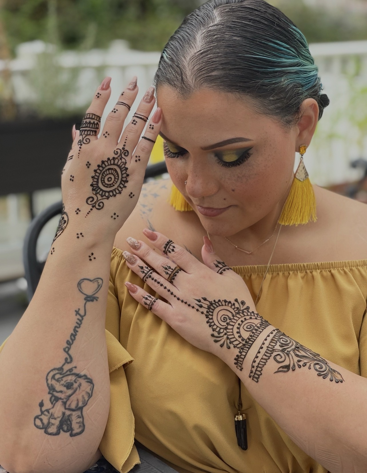 Woman with dark hair and yellow earrings, showing intricate henna designs on her hands and arms, wearing a yellow top.