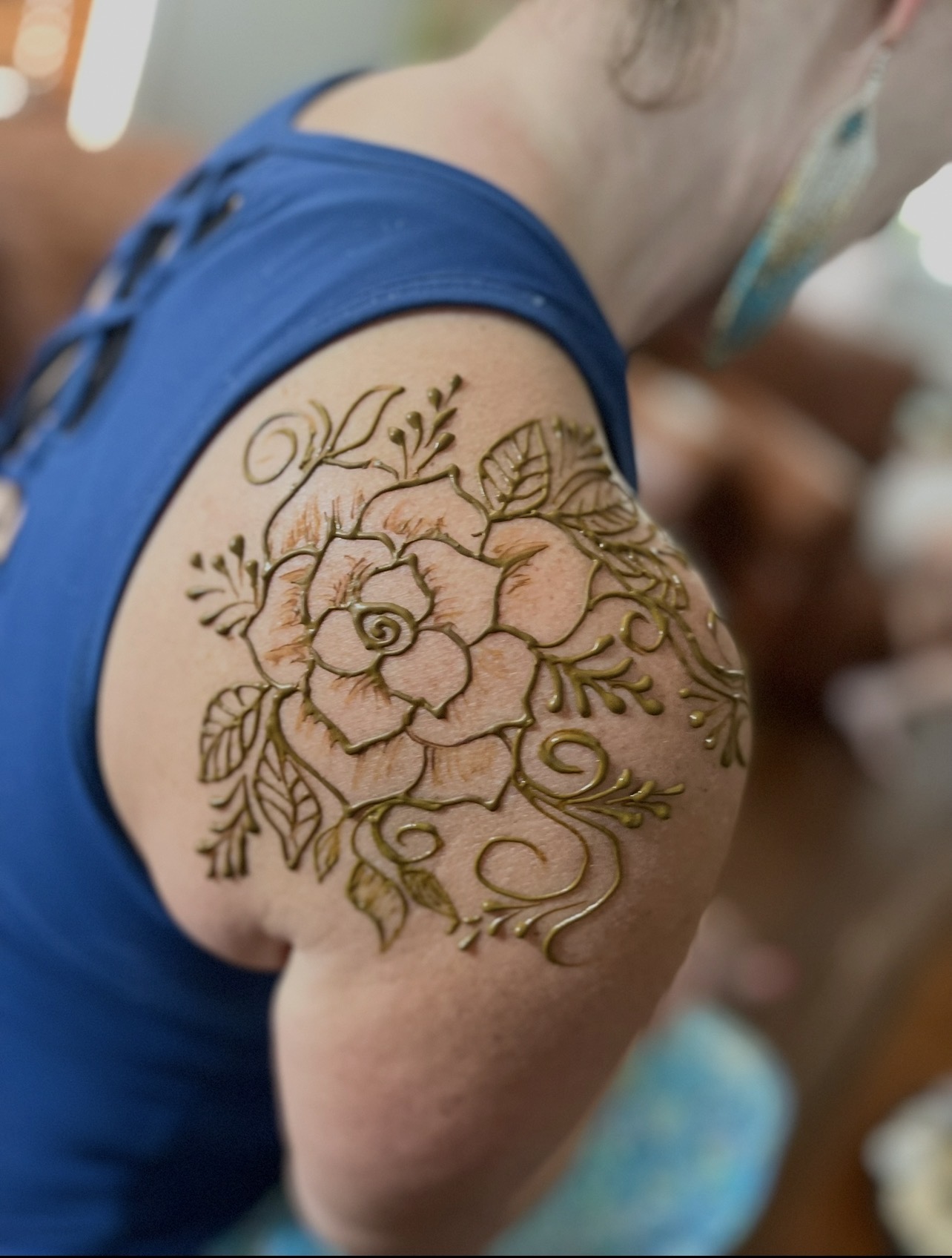 Close-up of a shoulder tattoo with a large flower, leaves, and decorative elements on a person wearing a blue top.