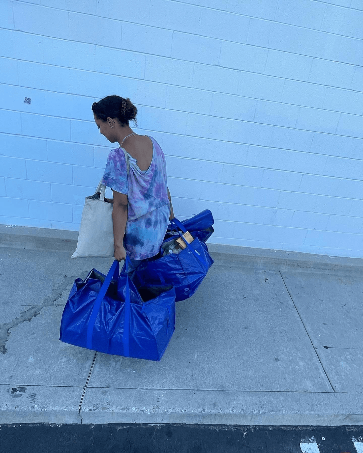 Woman with a tote bag and a large blue bag standing against a light blue wall, looking down, on a sidewalk.