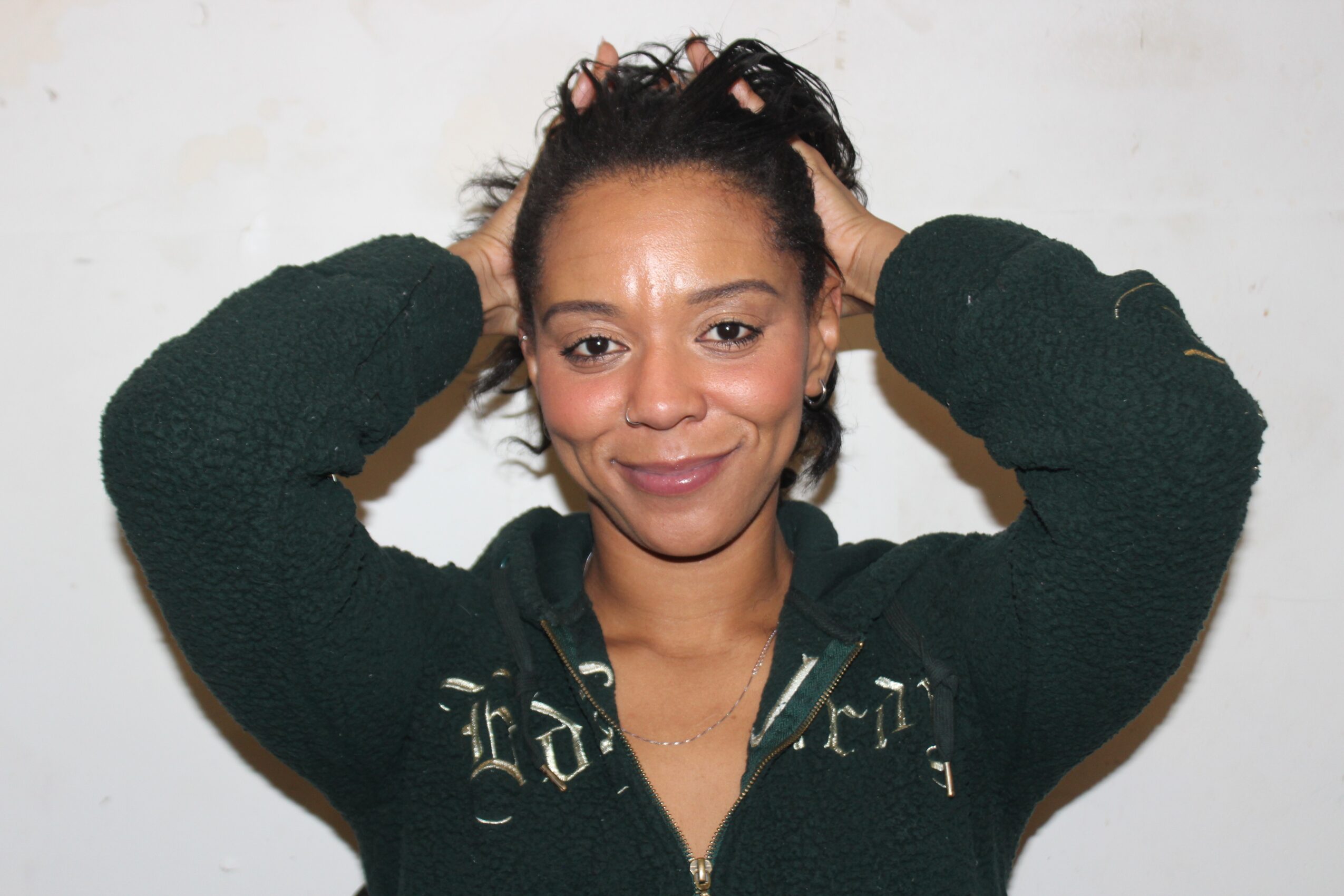 Woman with dark hair pulled back, smiling, hands on head, wearing a dark jacket, standing against a white background.