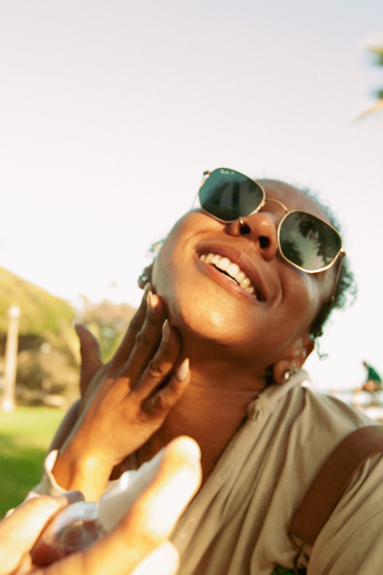 Woman smiling outdoors wearing sunglasses, touching her face, with a bright sky and greenery in the background.