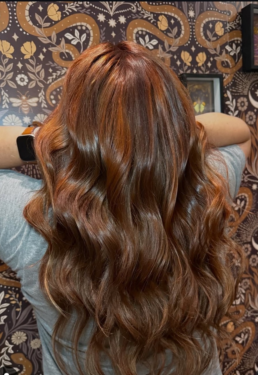 Back of a person with long, wavy, reddish-brown hair, in front of a patterned wallpaper background.