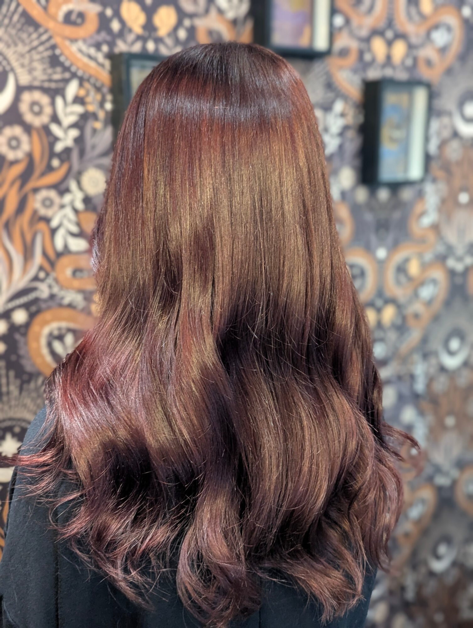 Back of a person with long, wavy, reddish-brown hair, standing in front of a patterned wall.