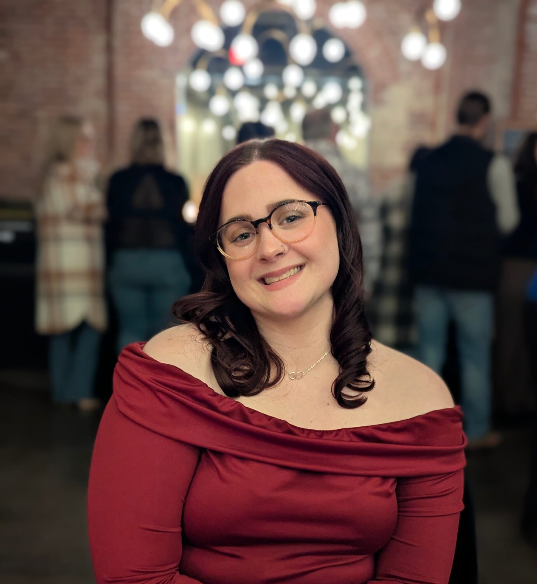 Smiling woman with glasses and dark hair, wearing a red off-shoulder top, in a festive indoor setting.