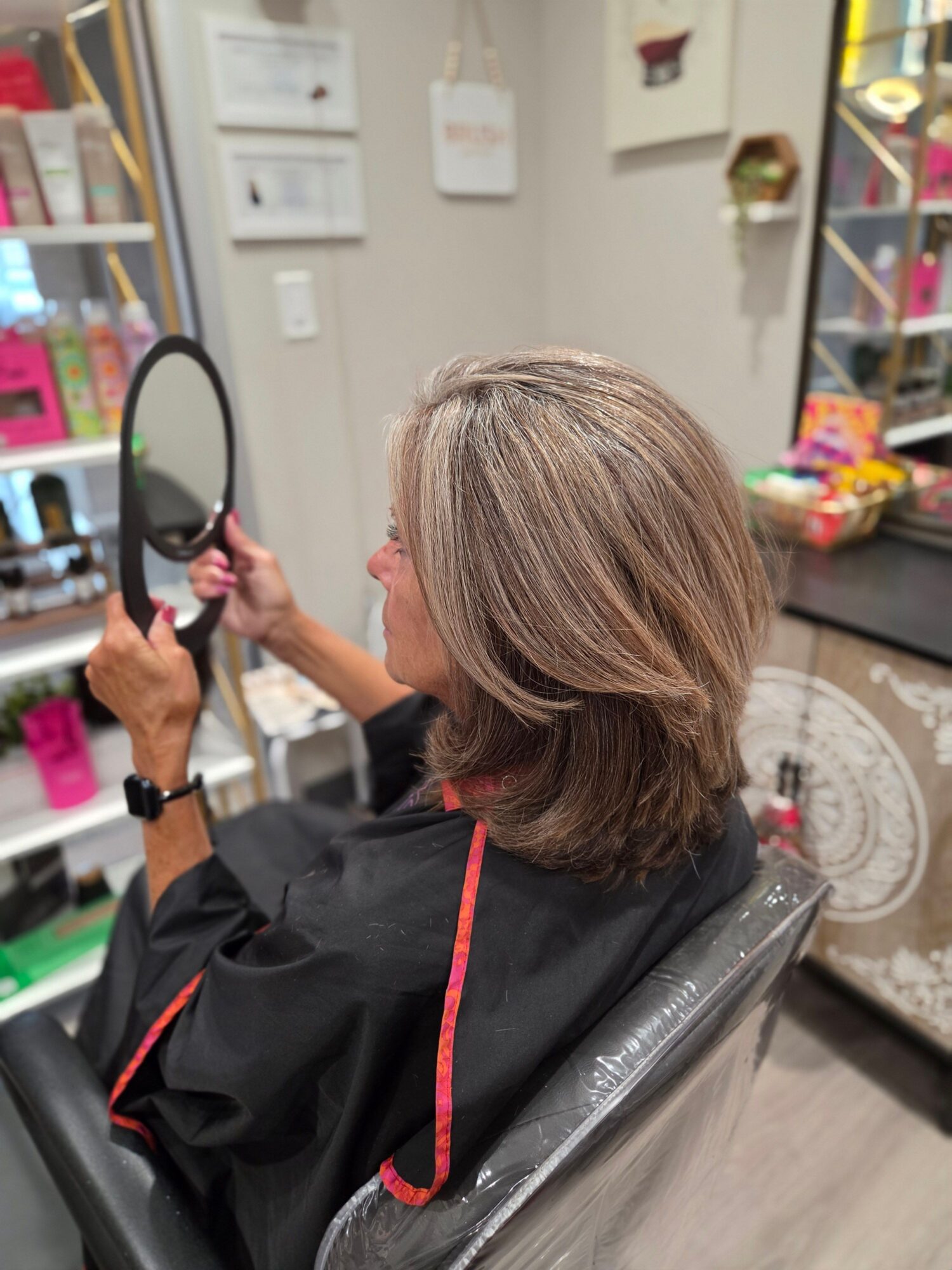 Person with shoulder-length hair sitting in a salon chair, holding a mirror, with salon equipment in the background.