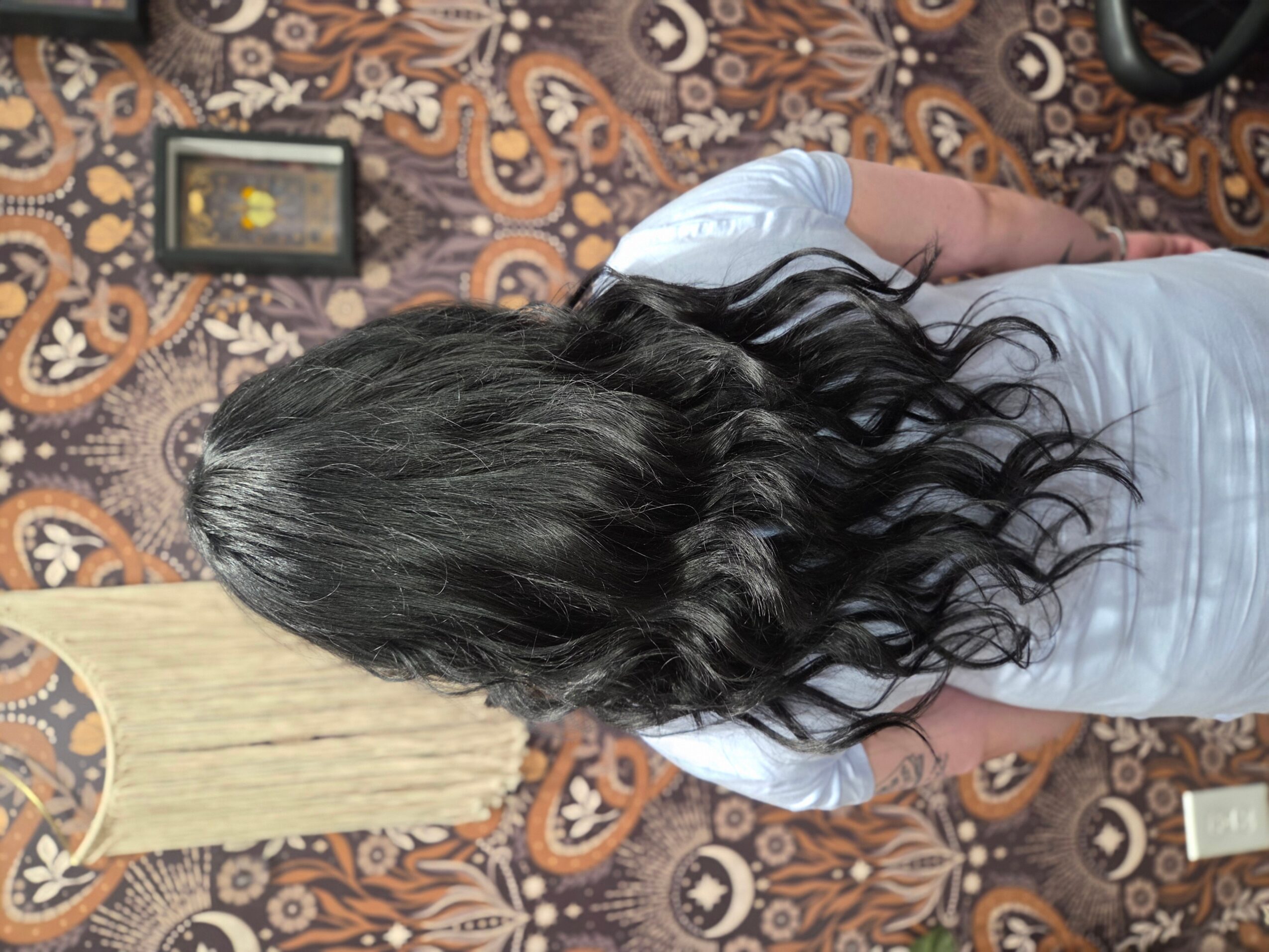 Person with long, wavy black hair standing on a patterned carpet, viewed from behind, wearing a light-colored shirt.