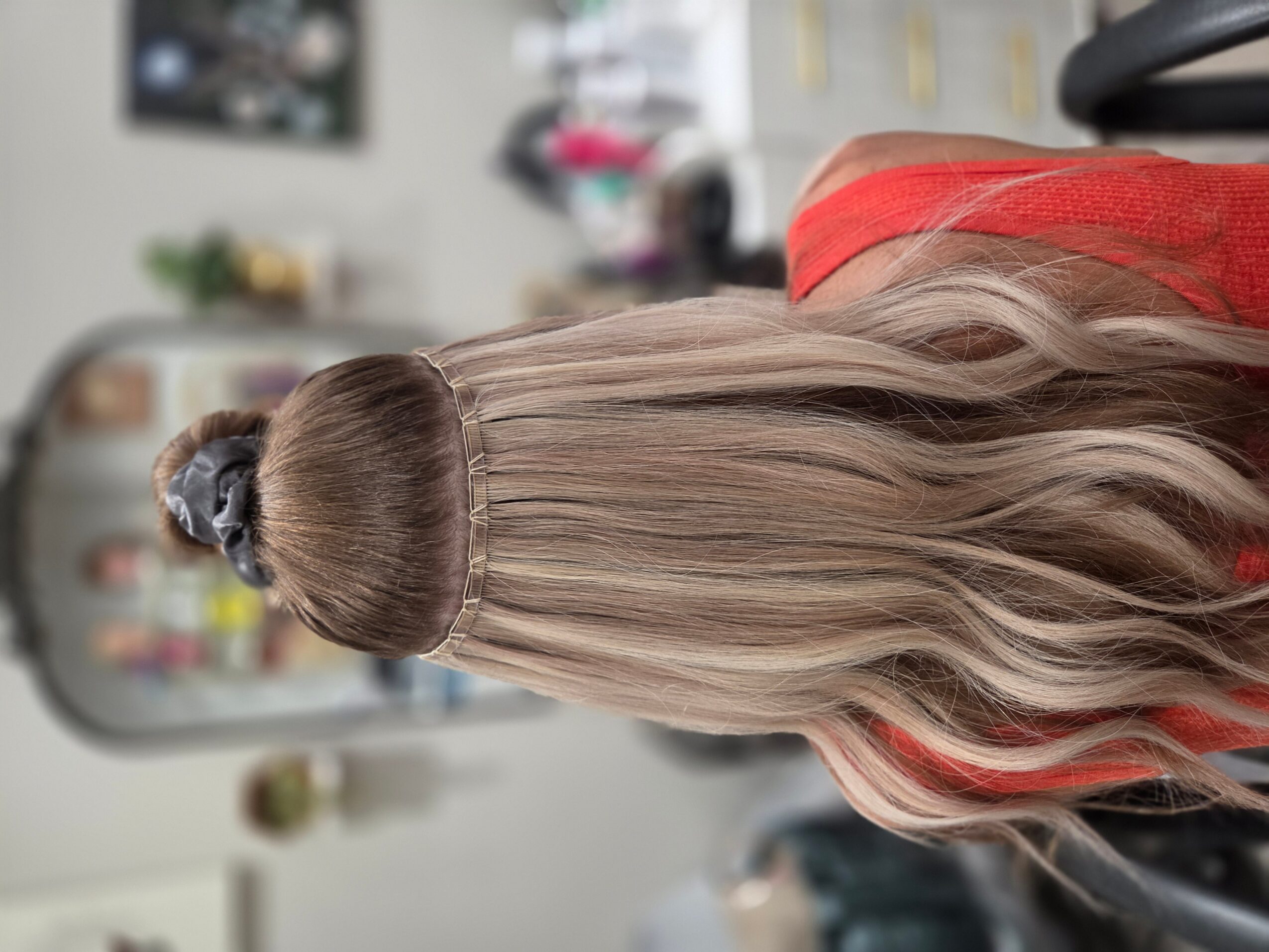 Person with long, wavy hair sitting in a salon chair, facing away, with a blurred background of salon equipment.