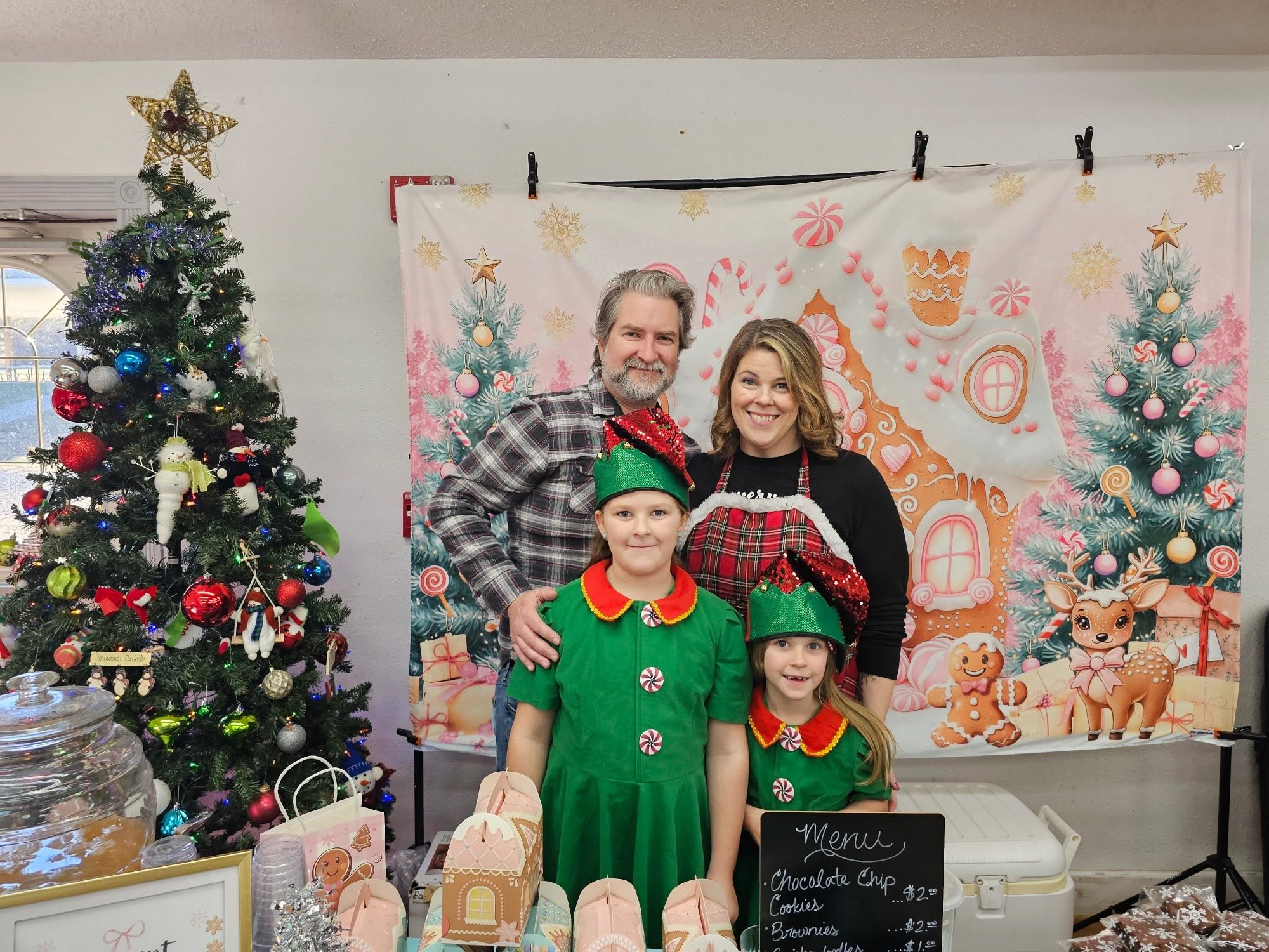 Family of four dressed in Christmas costumes standing in front of holiday backdrop with decorated Christmas trees.