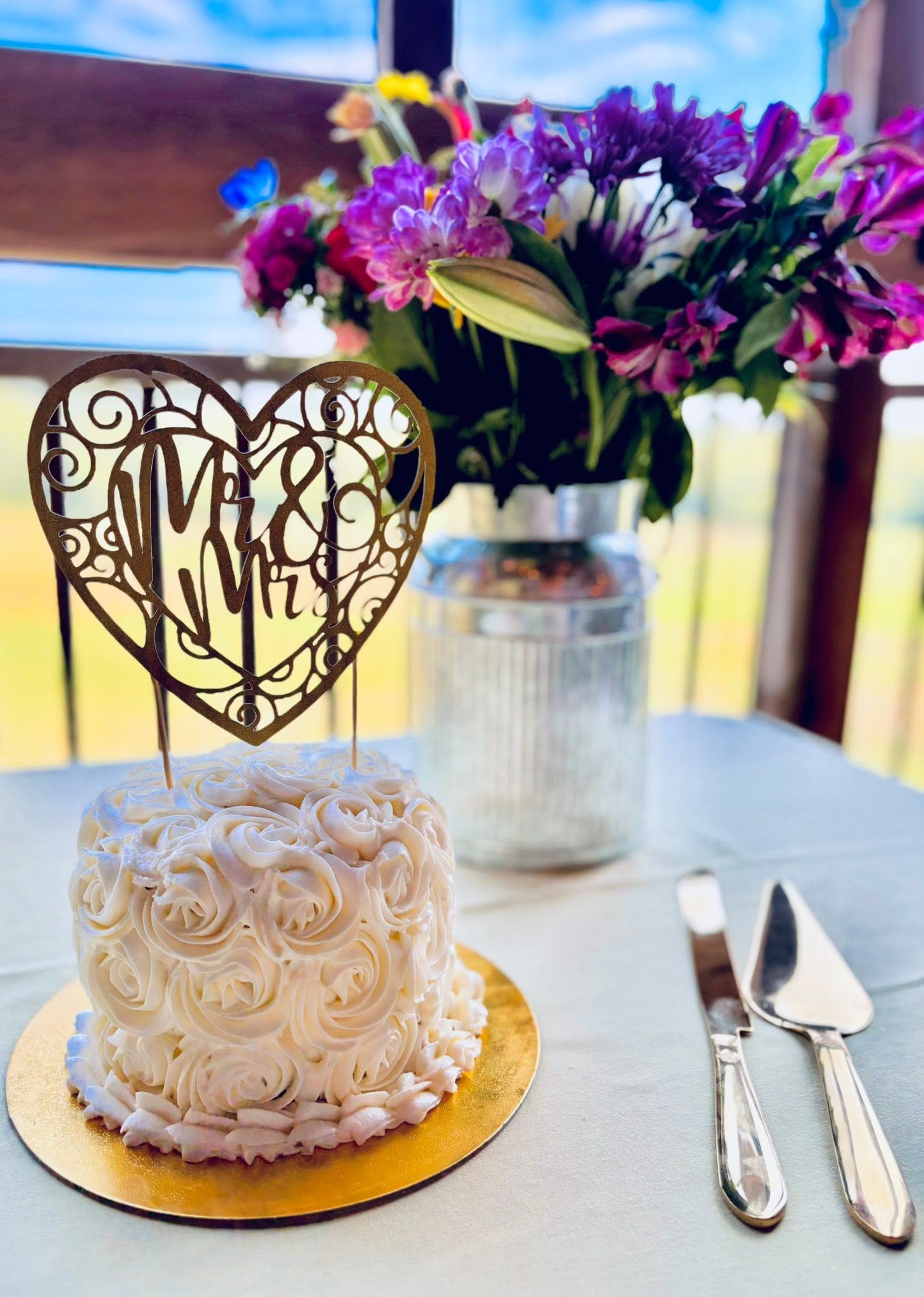 A small white cake with rosette decorations on a gold base, a heart-shaped topper, and a bouquet of purple and pink flowers in a jar behind it, on a table.