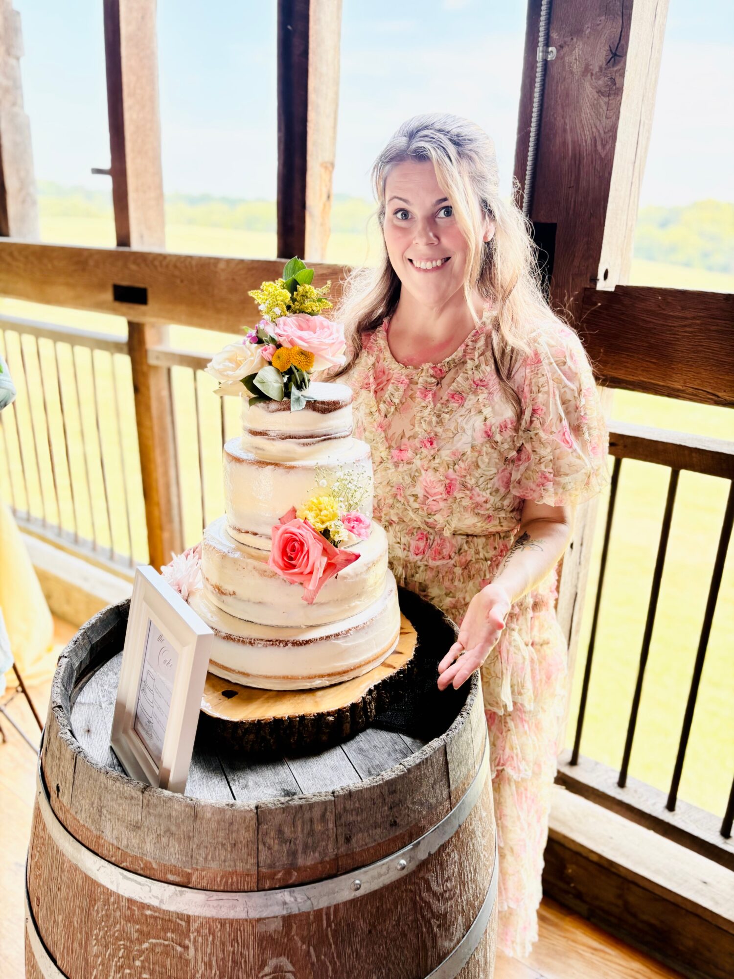 Woman with long blonde hair in floral dress smiling next to a three-tiered cake with flowers, on a wooden barrel, indoors.