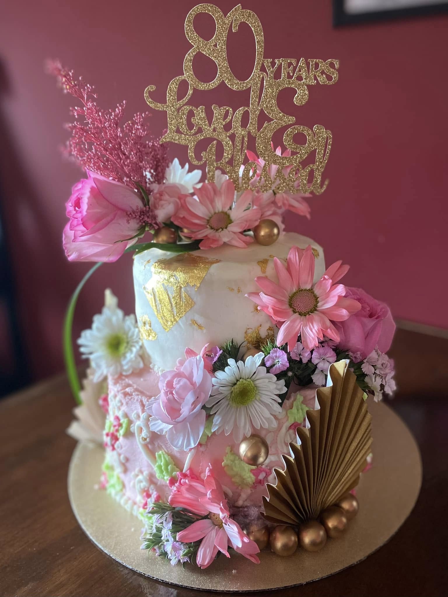Decorative cake with pink and white flowers, gold accents, and a gold topper reading '80 Years Love & Joy'.