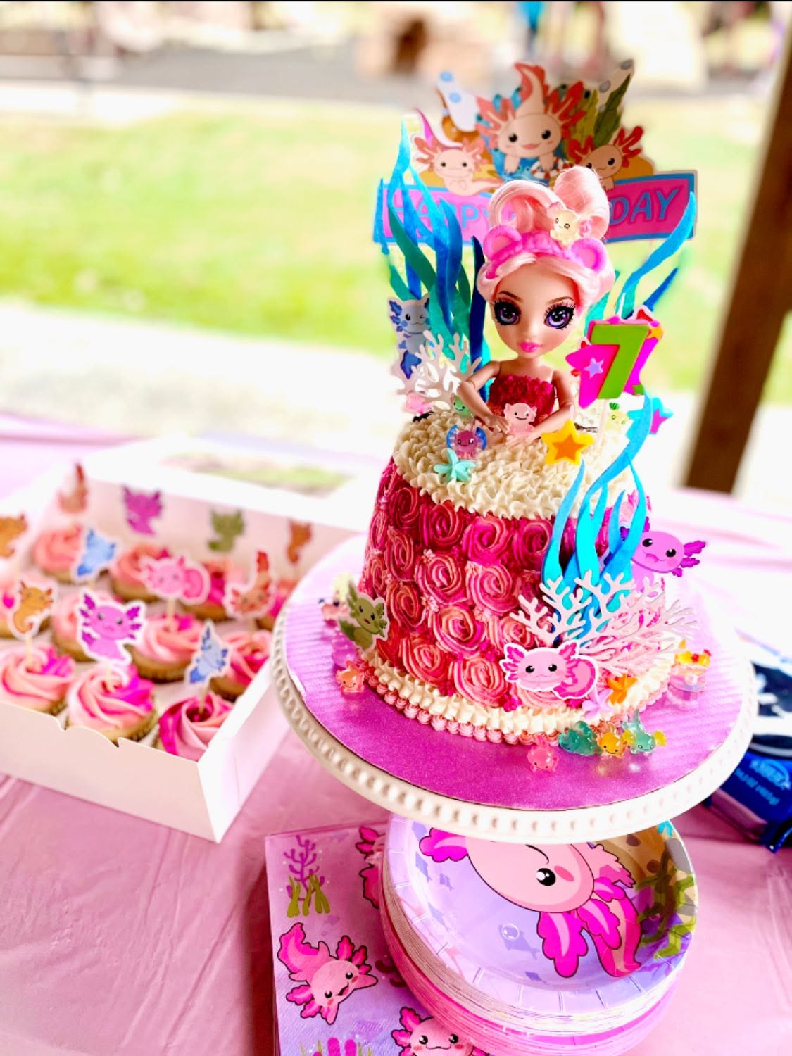 Colorful birthday cake with a doll figure, decorated with pink roses, blue flames, and animal figures, on a pink table.