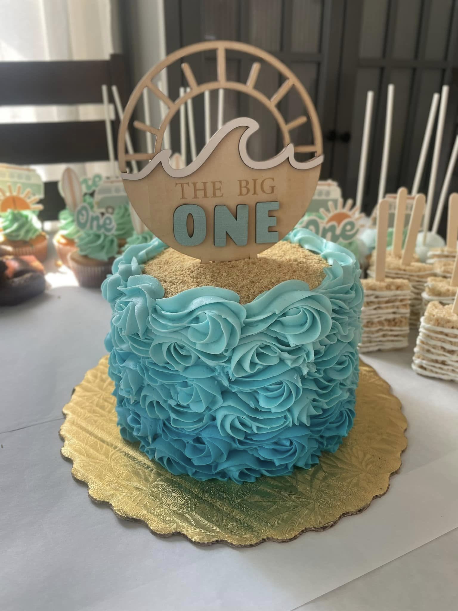 A round cake decorated with blue rosette frosting, topped with a wooden cake topper reading