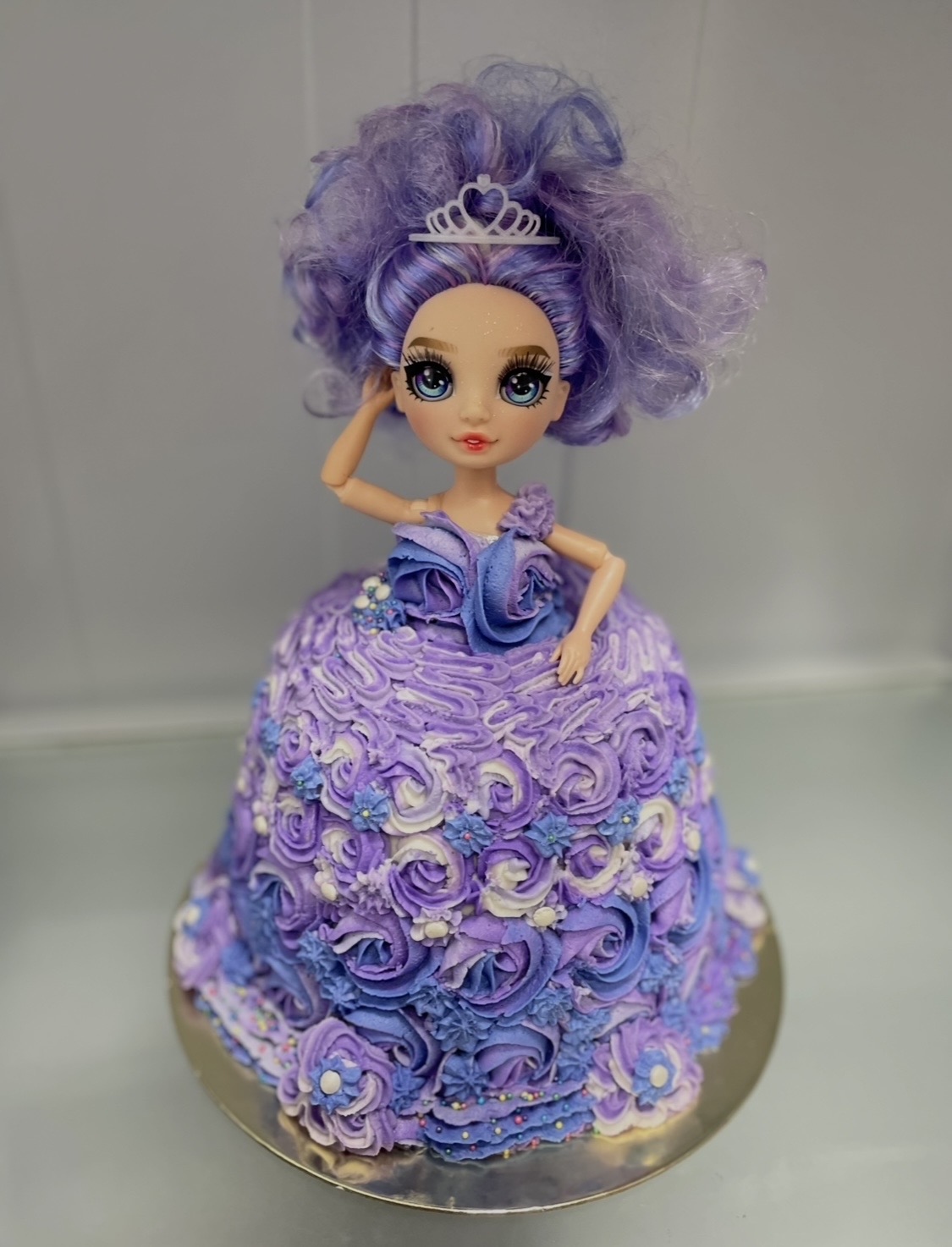 Doll with purple curly hair wearing a tiara and a purple dress with rosette decorations, standing on a cake.