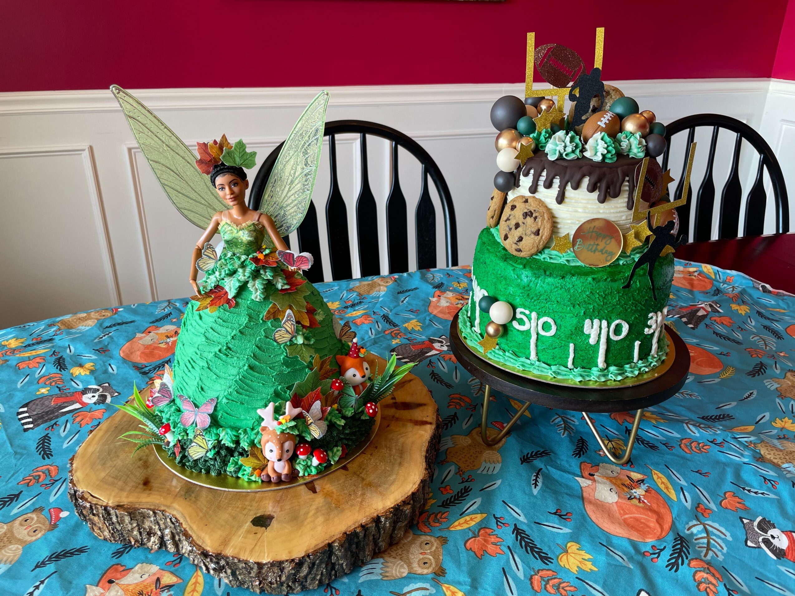 Fairy figure with green dress and wings, surrounded by small animals, on a wooden slab; decorated cake with green, brown, and gold elements on a table with a colorful tablecloth.