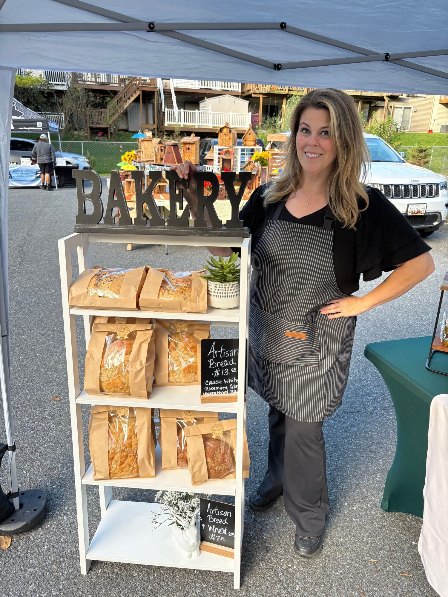 Woman standing next to a display of baked goods at an outdoor market, with tents and other stalls in the background.