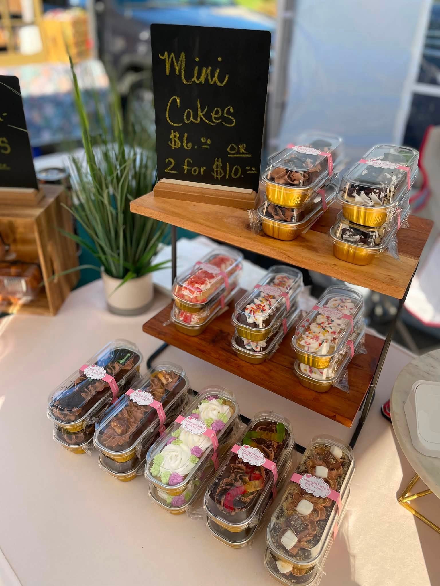 Mini cakes displayed on a two-tier wooden stand with a black chalkboard sign, in a store setting.