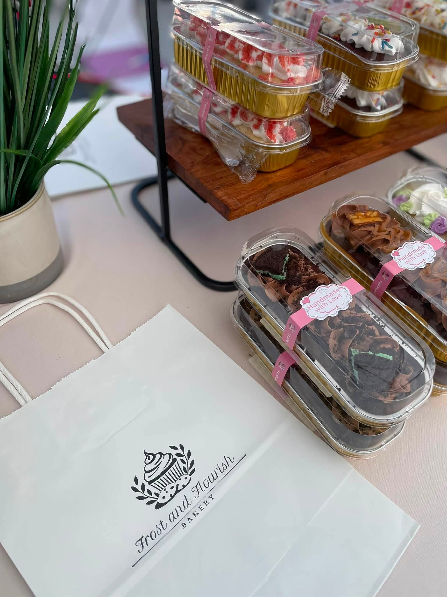 Clear plastic containers with baked goods on a table, a white paper bag with a logo, and a wooden shelf with more treats.