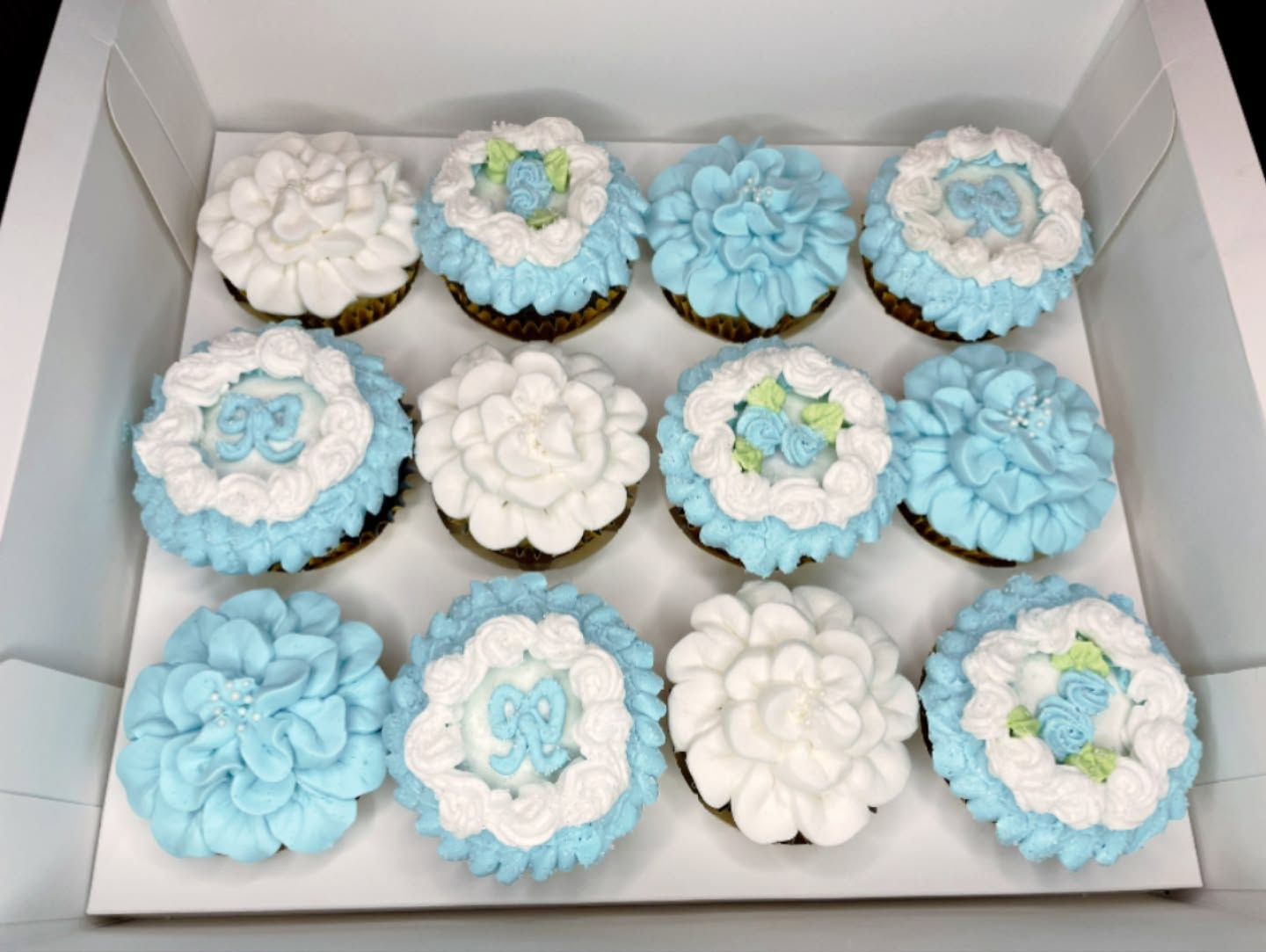 Box of twelve cupcakes decorated with white and blue floral icing, some with butterfly designs, arranged in three rows.
