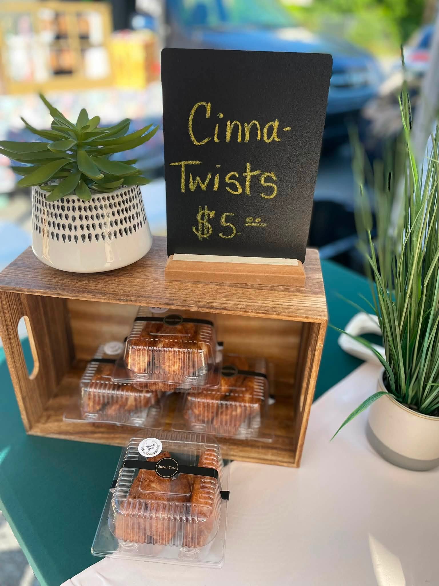 Display of cinnamon twists in clear containers on a wooden stand, with a black sign reading 'Cinna-Twists $5', surrounded by plants.